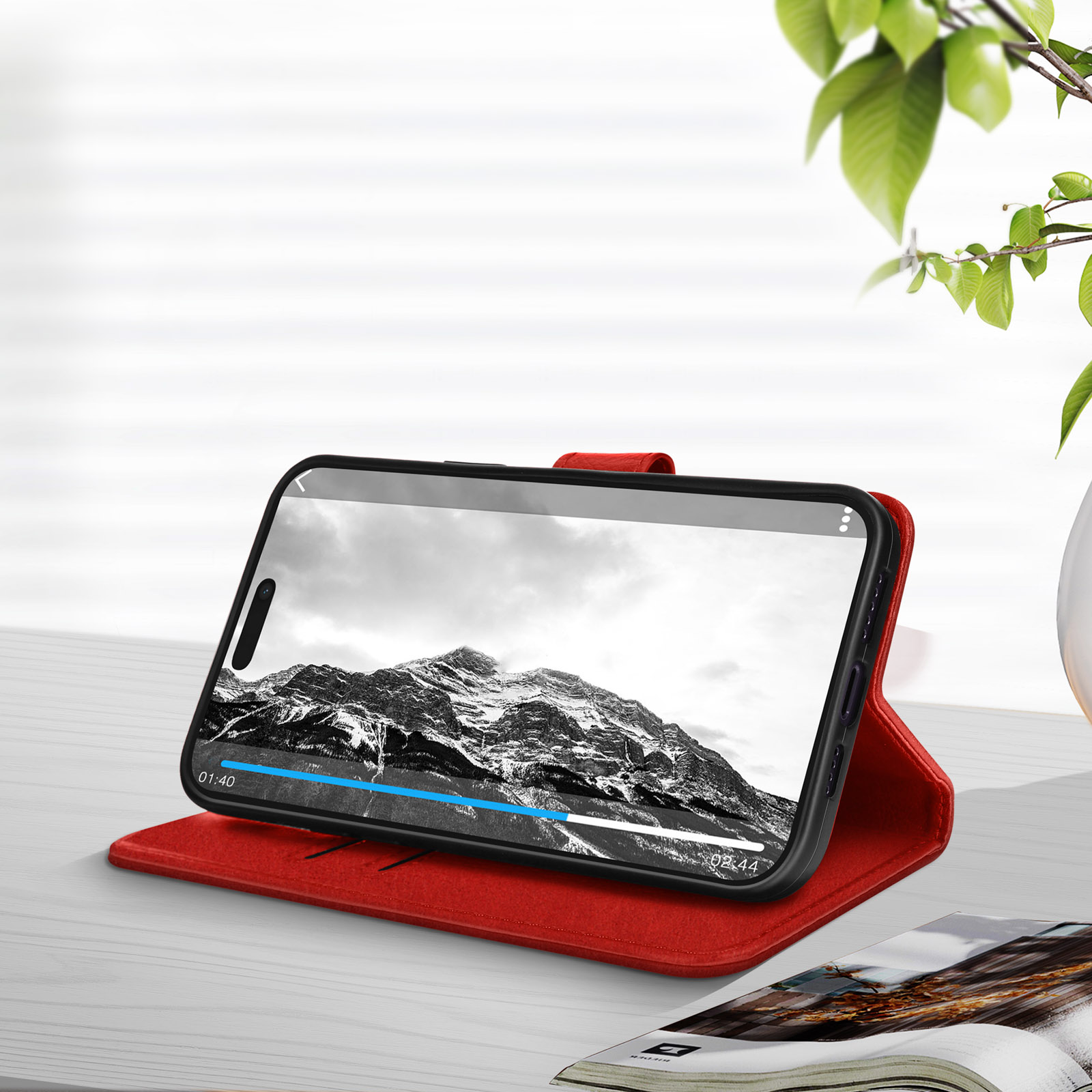 Téléphone dans étui rouge, écran montrant vidéo. Plante et livre en arrière-plan.