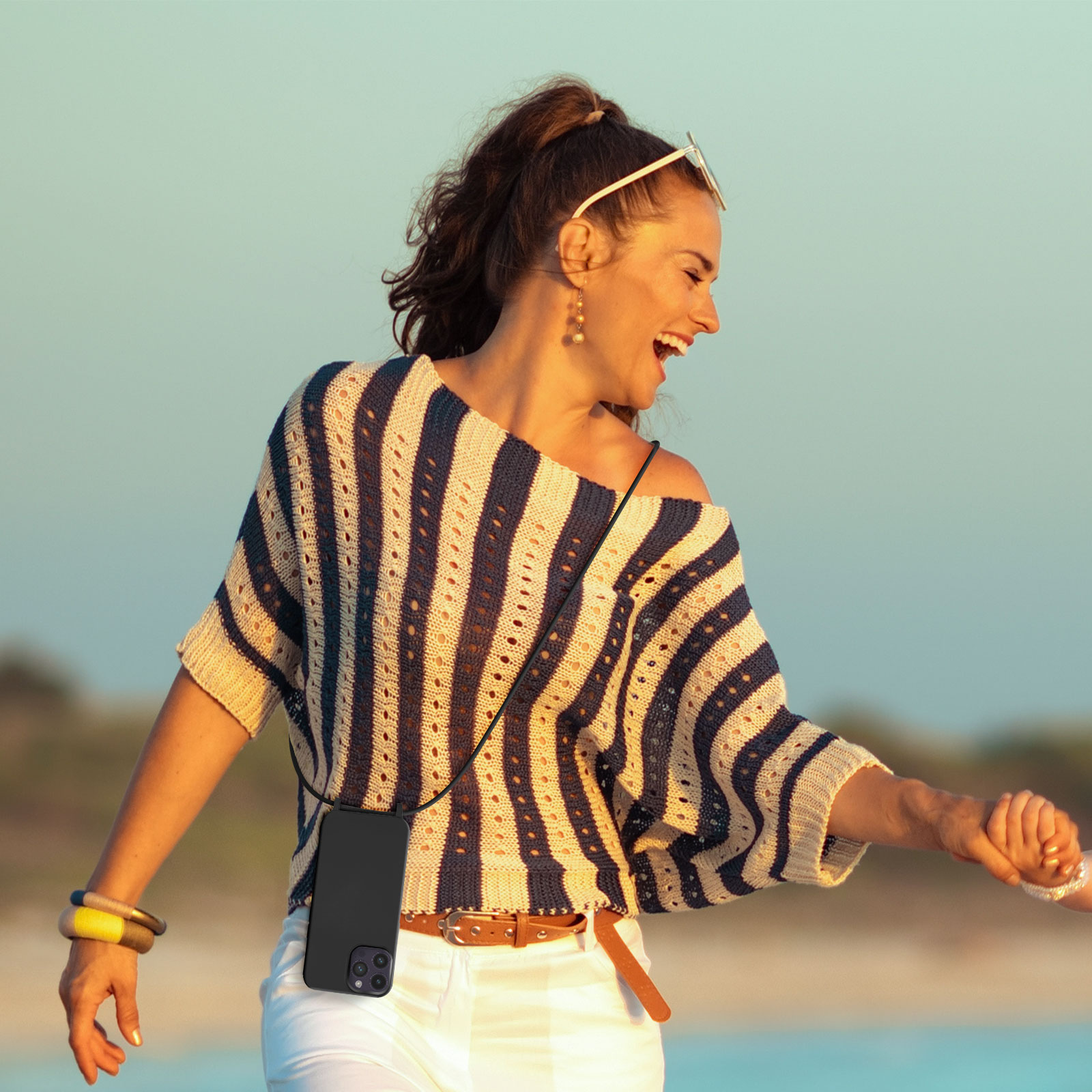 Vrouw met een telefoon aan een koord, lachend en genietend van de zon. Achtergrond: strand.
