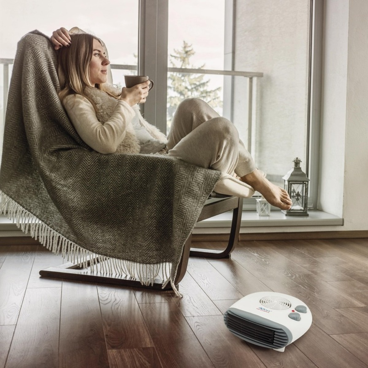 Una mujer sentada en una silla con una manta, sosteniendo una taza cerca de un calefactor.