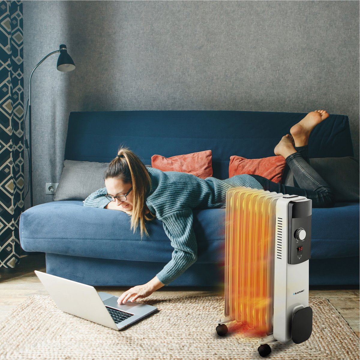 Frau benutzt Laptop neben Ölradiator. Liegt auf blauem Sofa. Rote Decke. Schwarzer und grauer Heizkörper.