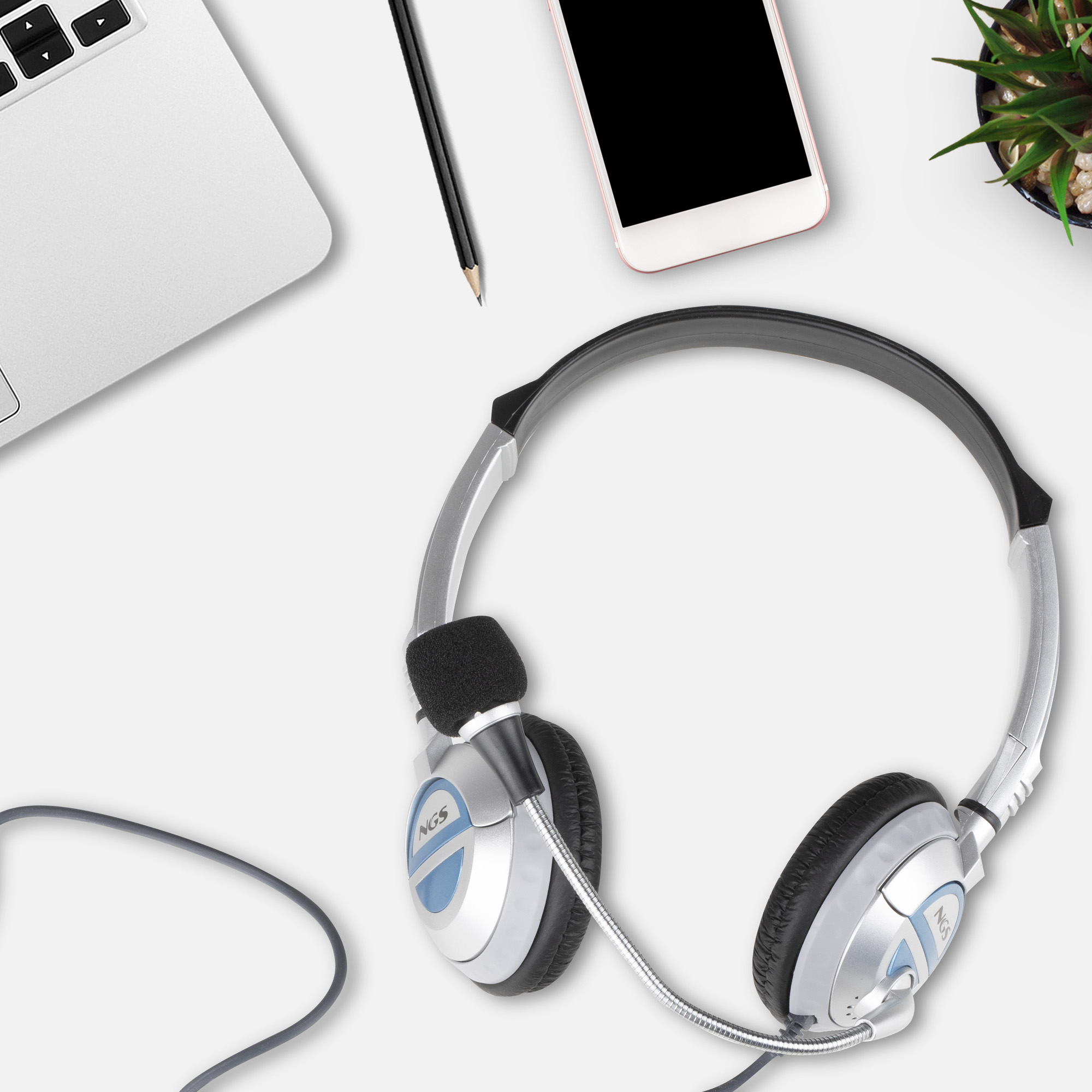 Vista cenital de un escritorio. Auriculares, portátil, teléfono, lápiz y planta sobre una superficie blanca.