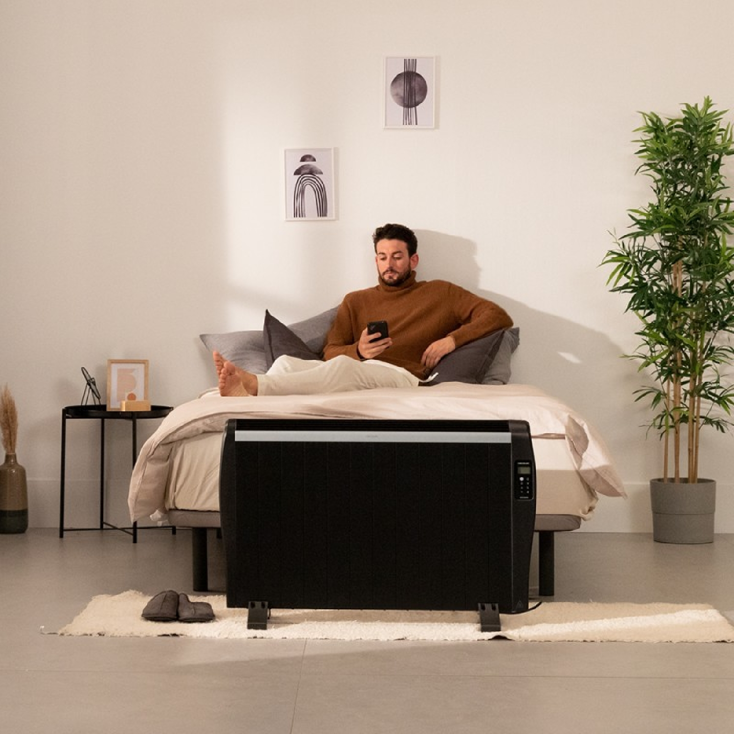 Mężczyzna używa telefonu w łóżku. Czarny grzejnik przed łóżkiem. Dwa obrazy na ścianie.