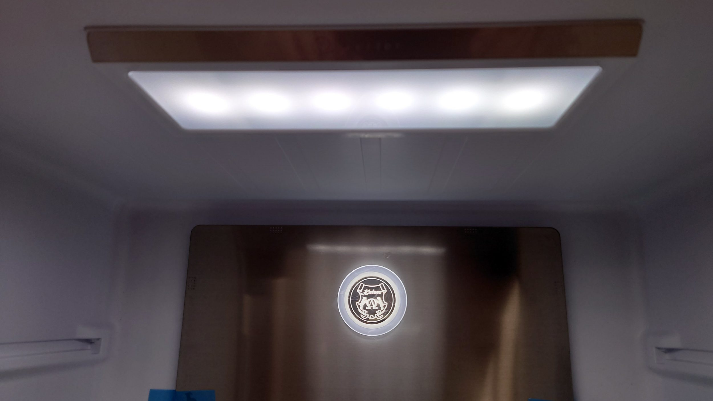 Im Inneren eines Kühlschranks. LED-Lichtfeld oben. Edelstahlrückwand mit kreisförmigem Logo sichtbar.