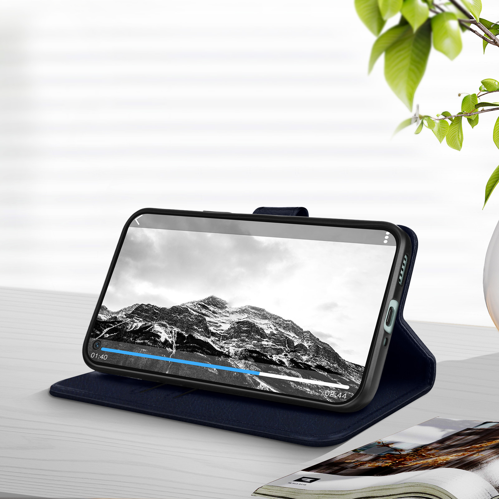 Ein Smartphone, das ein Berglandschaftsvideo auf einem blauen Cover anzeigt, geöffnet auf einem Tisch neben einer Zeitschrift.