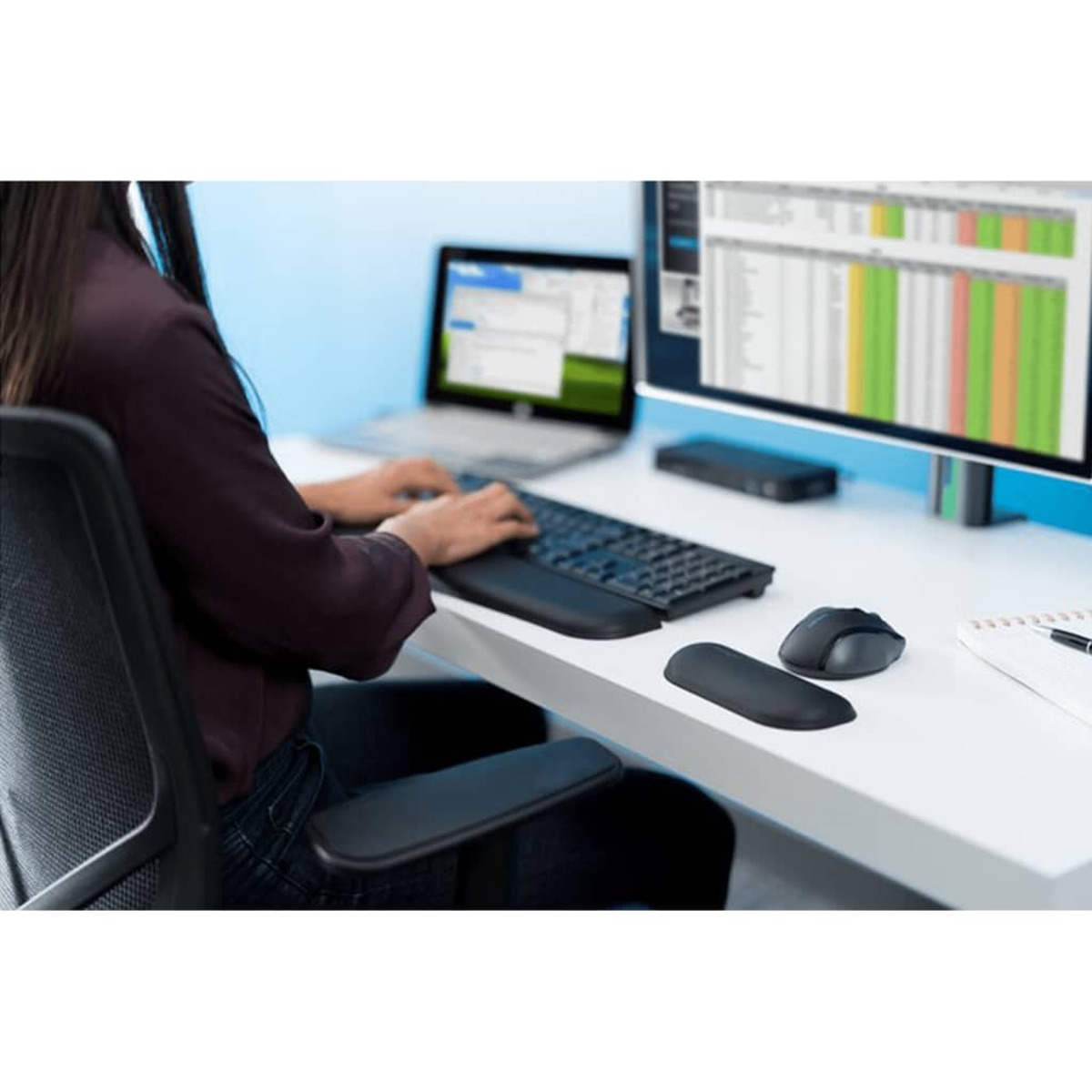 Una persona escribe en un teclado en un escritorio. Una computadora portátil y un monitor muestran datos.