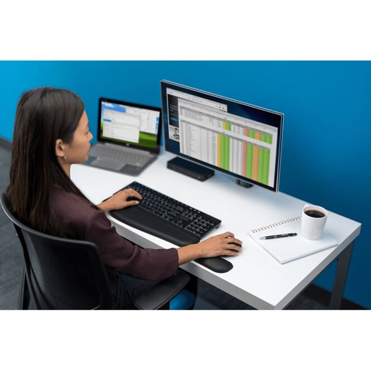 Una mujer está trabajando en un escritorio. Está sentada en un escritorio con una computadora y una computadora portátil. Hay una taza de café.