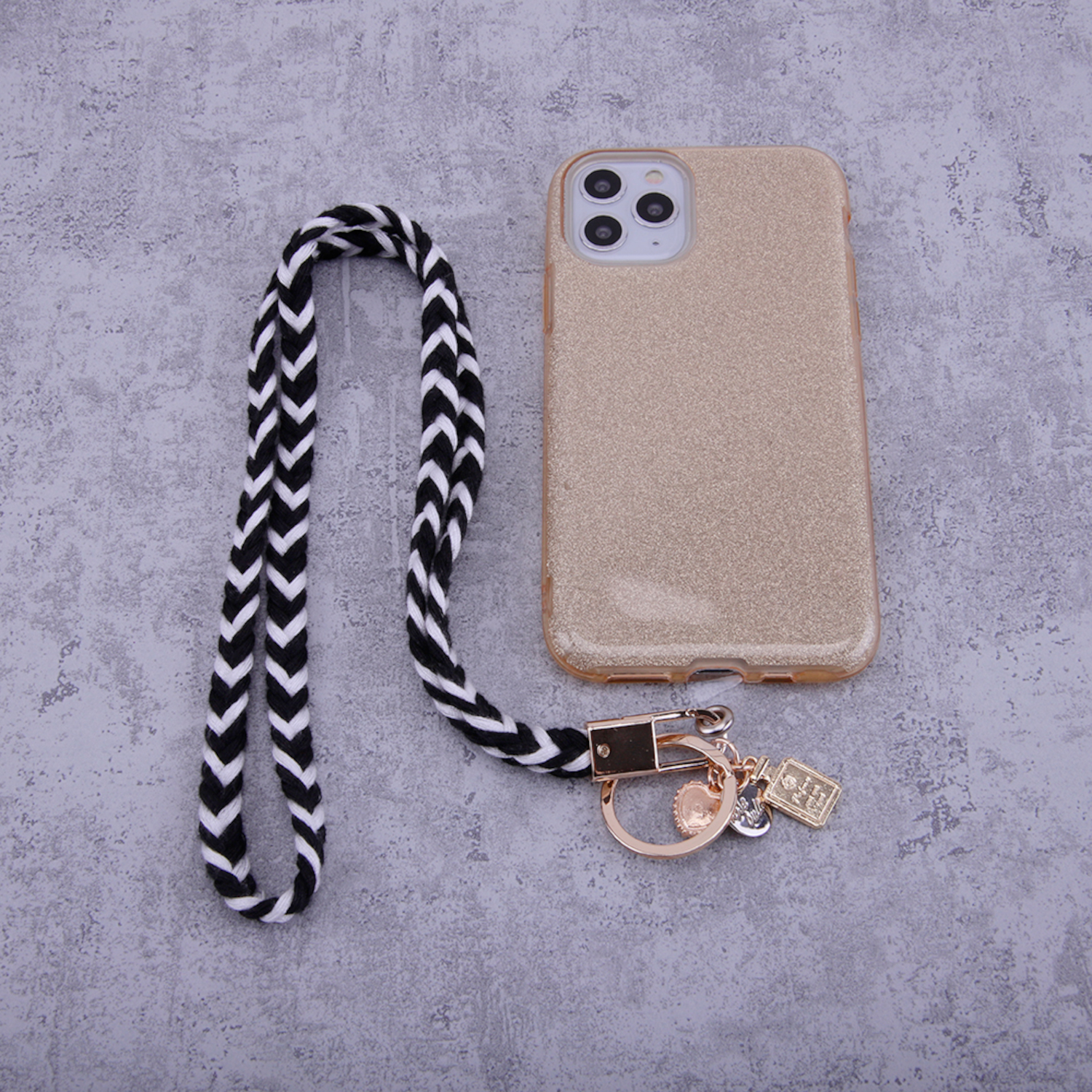 Goldenes Handycase, schwarz-weißes Schlüsselband, mit Ring und Anhängern auf grauem Untergrund.