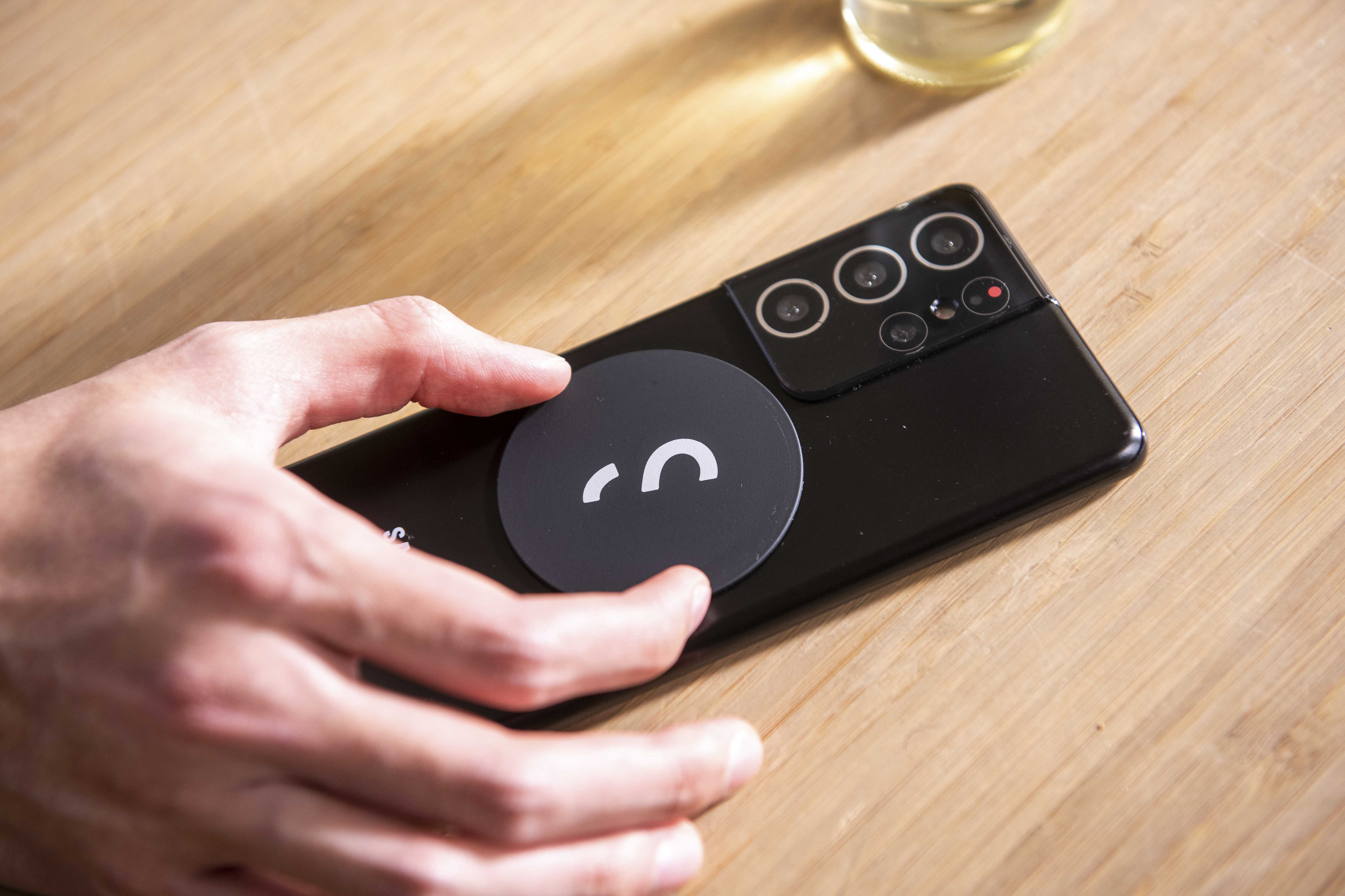 Hand hält schwarzes Telefon mit rundem schwarzem Zubehör auf einem Holztisch.
