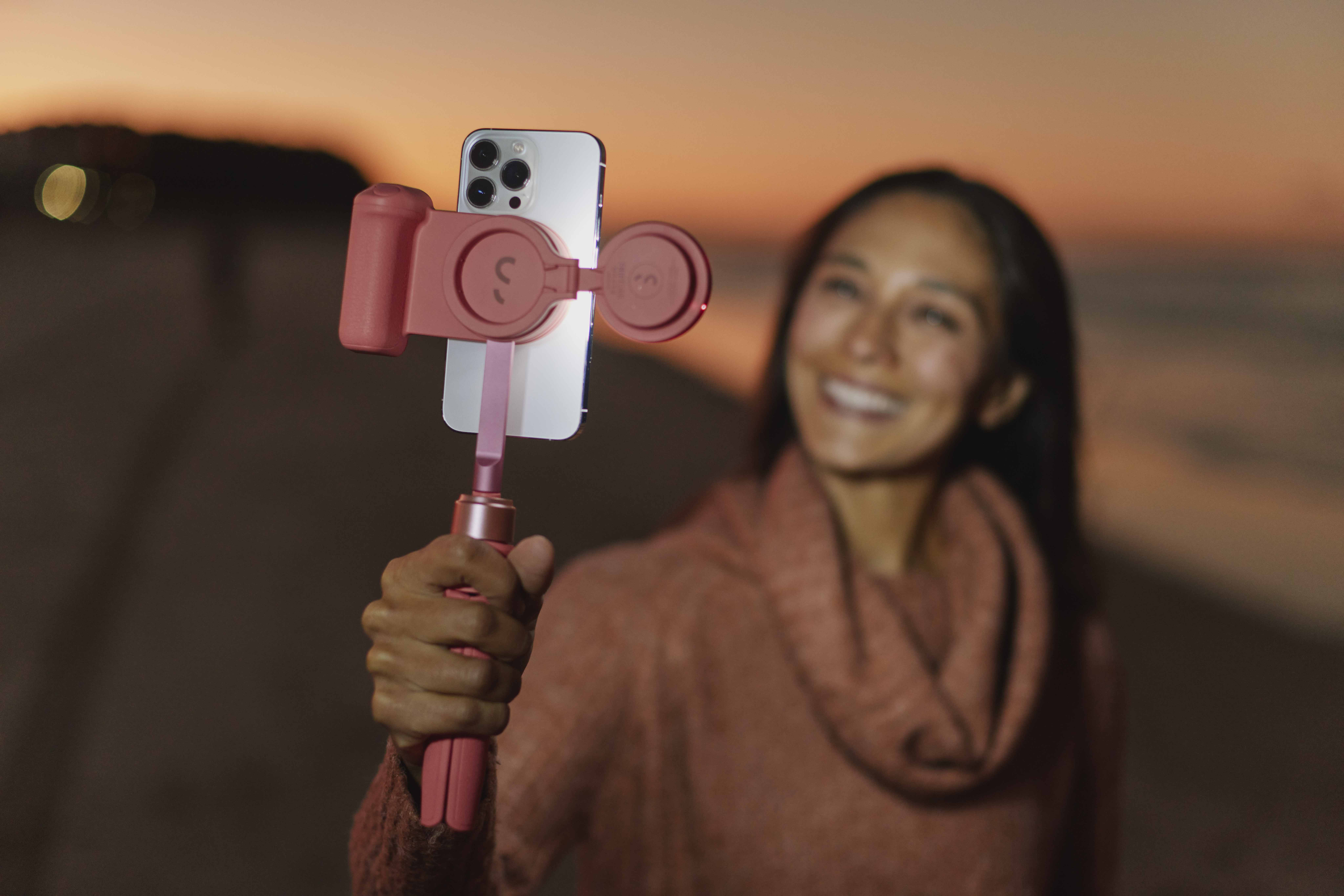 Eine Frau hält ein pinkfarbenes Telefon mit einem pinkfarbenen Selfie-Stick. Der Hintergrund zeigt einen Sonnenuntergang.