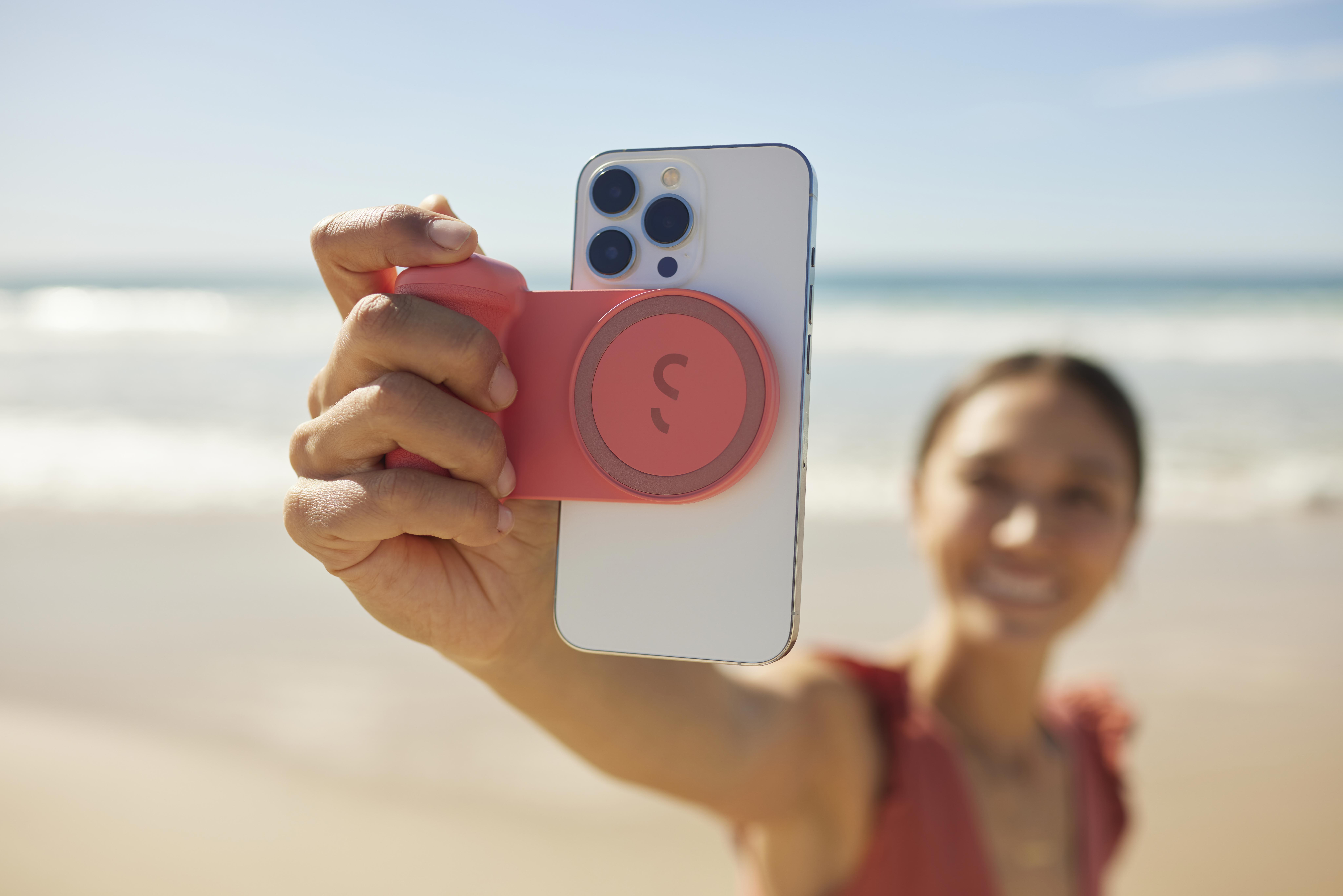Frau hält Telefon mit rosa Kameraaufsatz am Strand. Blaues Meer und Himmel.
