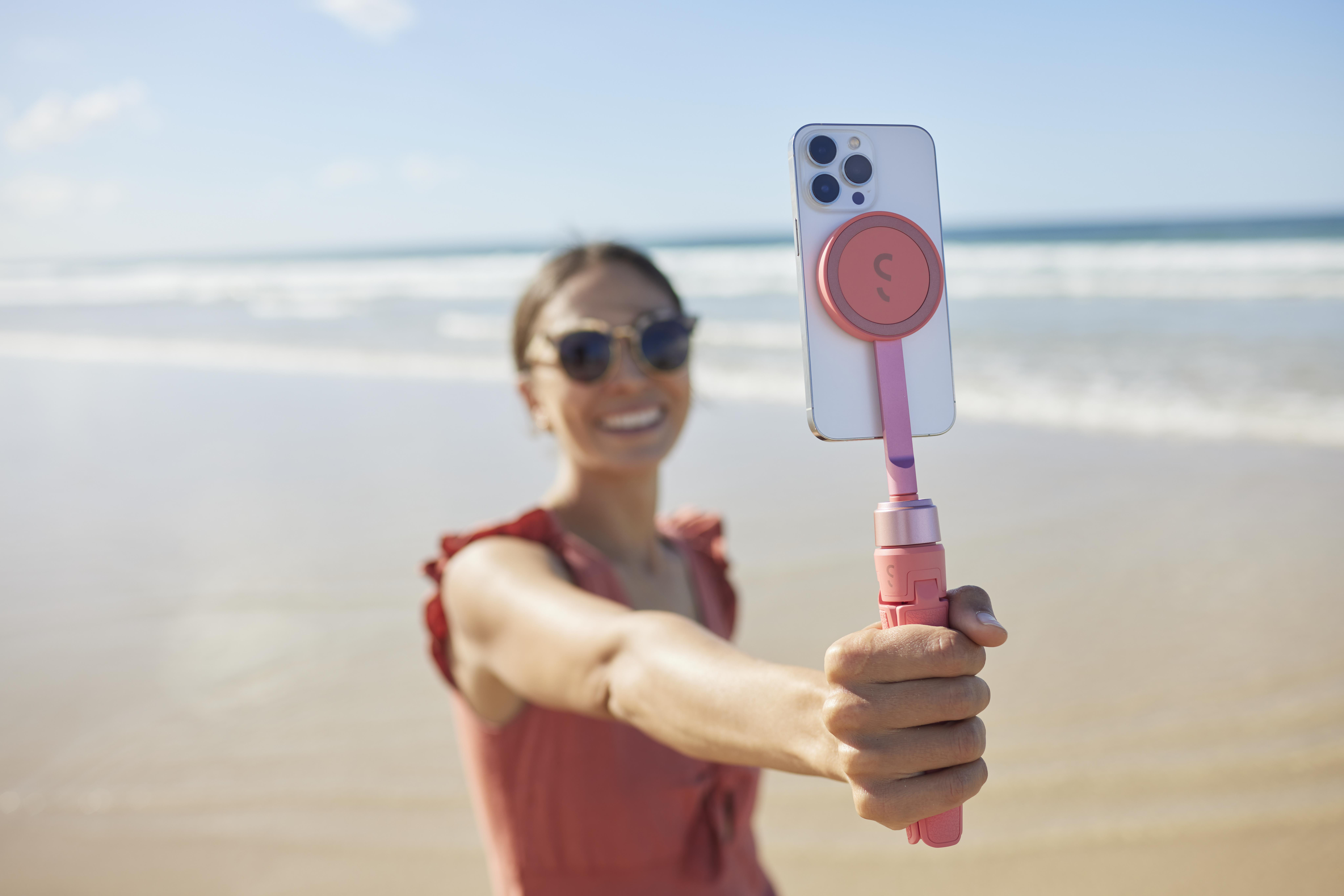 Eine Frau macht ein Selfie am Strand mit einem rosa Selfie-Stick. Im Hintergrund ist das Meer zu sehen.