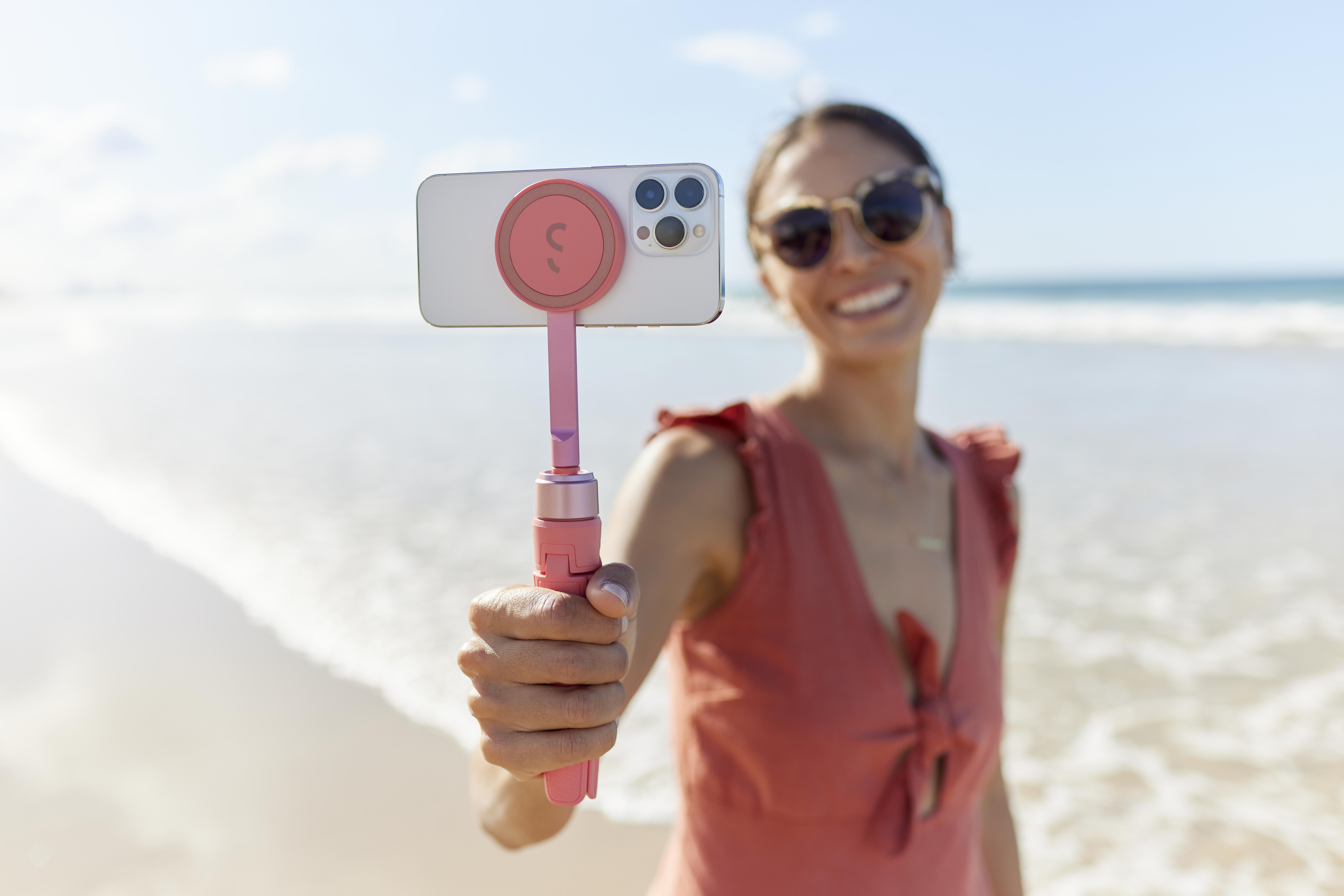 Eine Frau hält ein Telefon an einem pinkfarbenen Selfie-Stick, lächelnd am Strand.