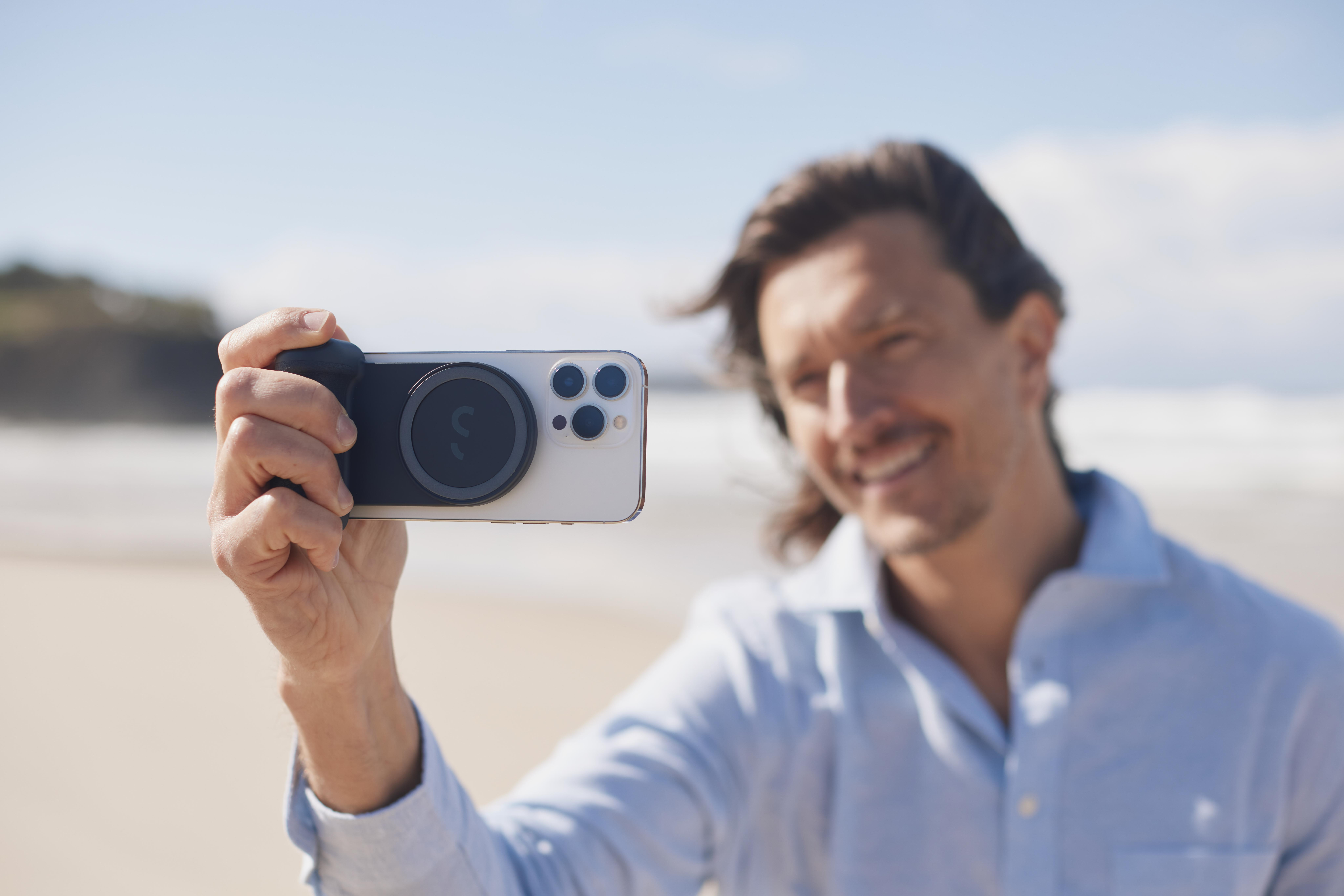 Mann hält Smartphone mit angesetzter Kameralinse, lächelnd. Strand und Ozean im Hintergrund.