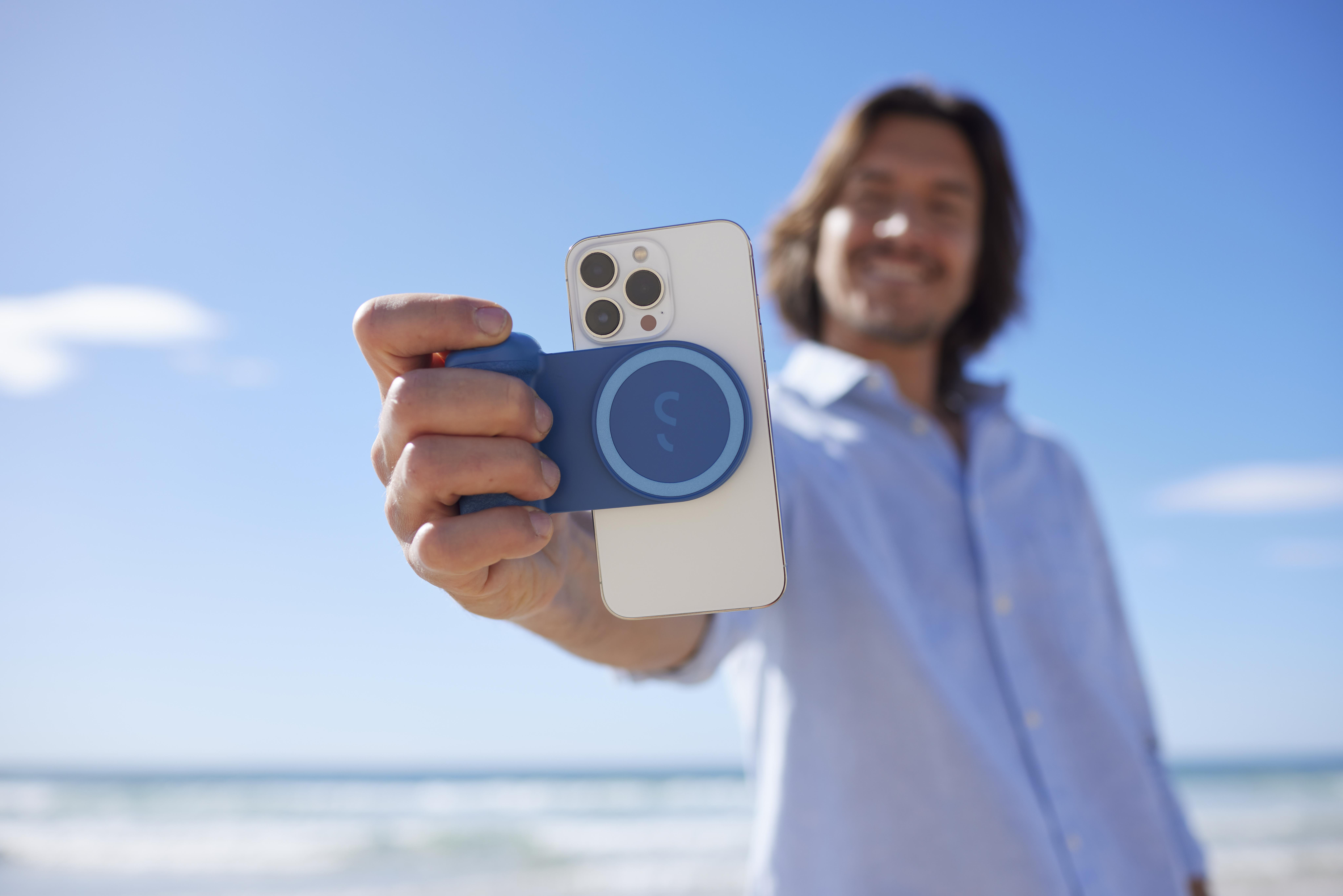 Mann hält ein weißes Telefon mit blauem magnetischem Zubehör. Hintergrund ist Strand mit blauem Himmel.
