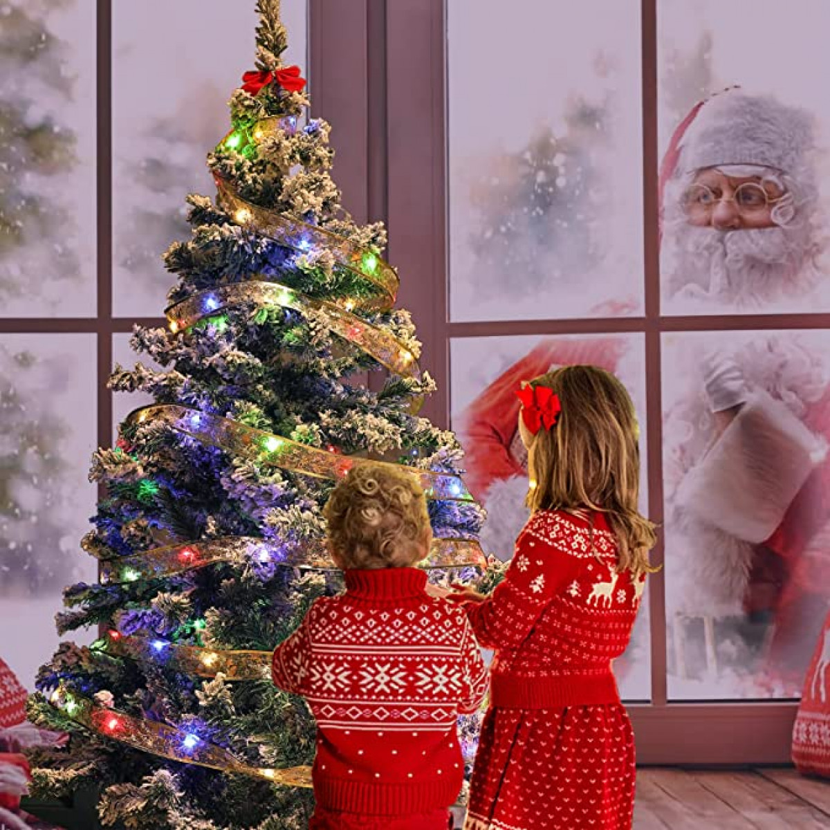 Zwei Kinder in roten Pullovern schmücken einen Weihnachtsbaum, der Weihnachtsmann ist im Hintergrund.
