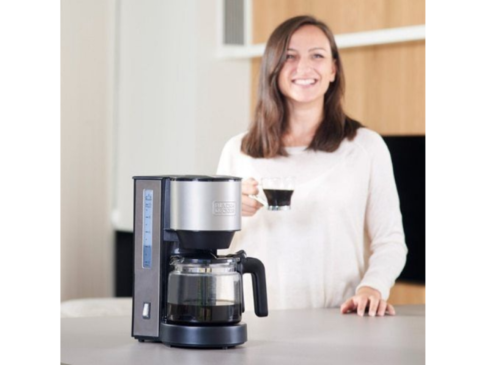 Kobieta uśmiecha się trzymając filiżankę kawy obok ekspresu do kawy na blacie.
