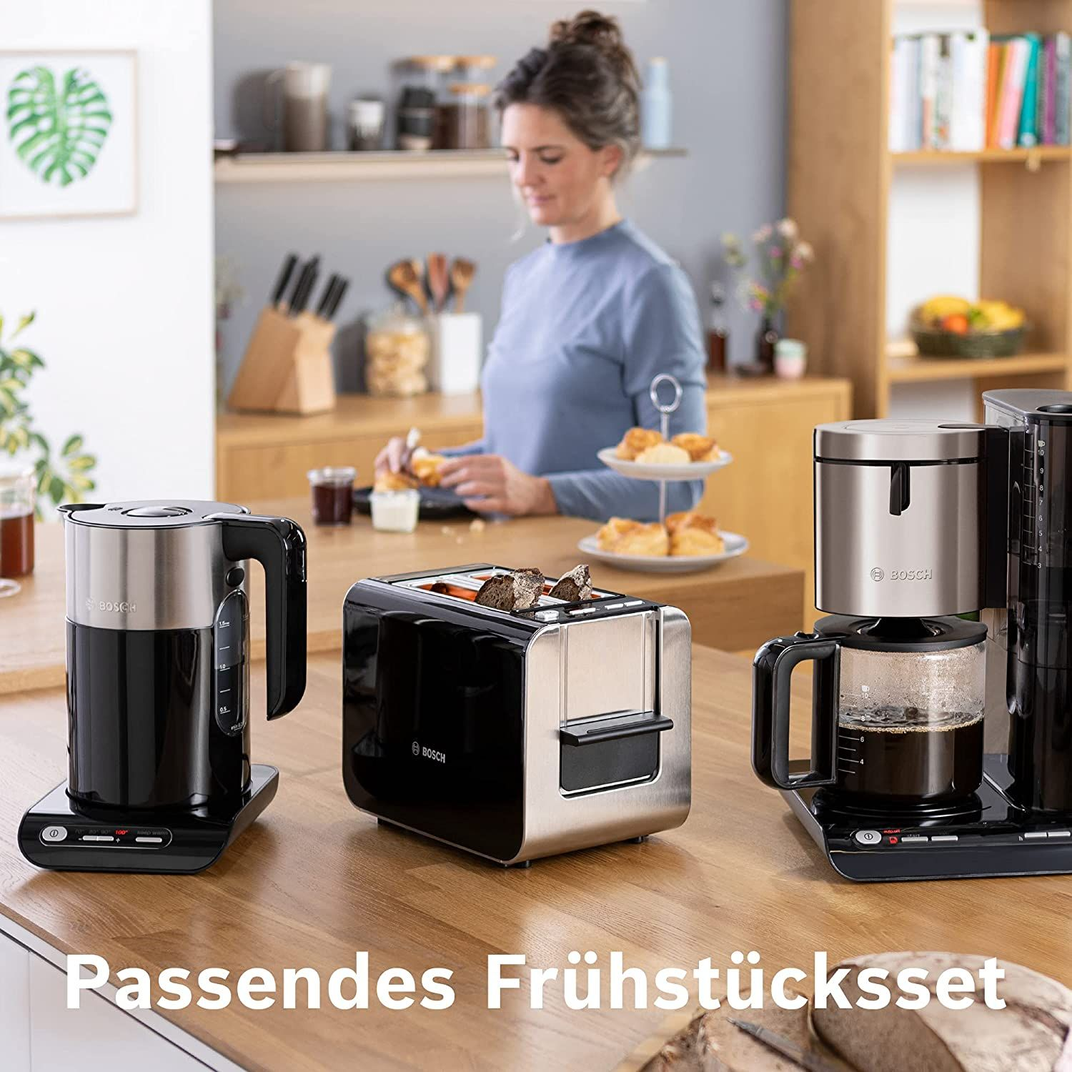 Elettrodomestici da cucina su un bancone di legno: bollitore, tostapane, macchina per il caffè e una donna sullo sfondo.