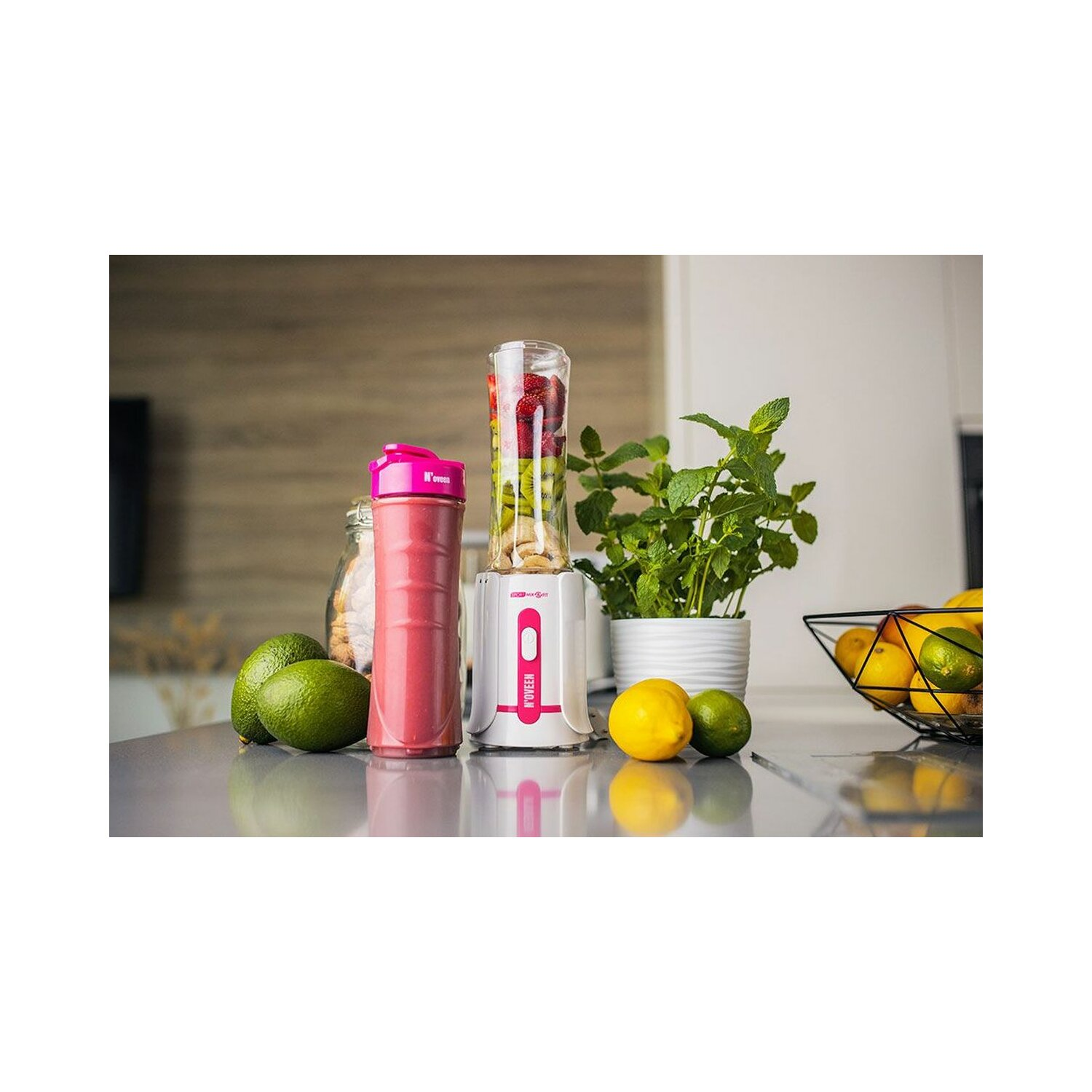 Blender z owocami i różową butelką, na białym tle, obecna jest również roślina, cytryny i limonki.