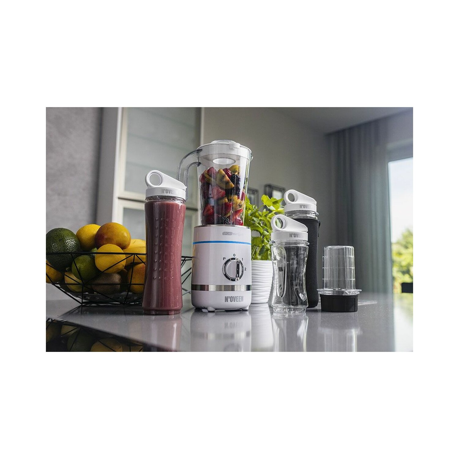 Blender z butelką smoothie i innymi butelkami. Obok miska owoców.