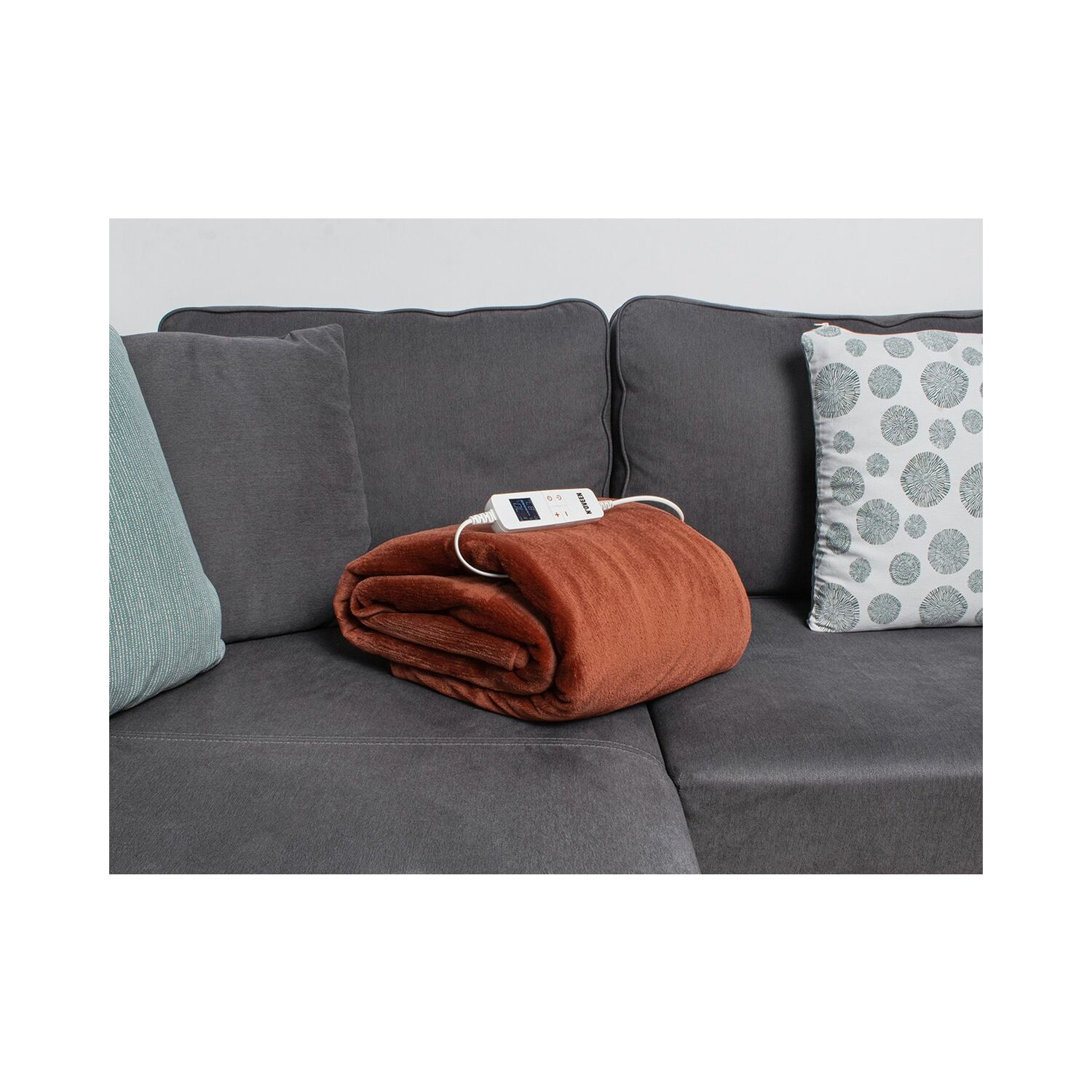 Braune Heizdecke auf grauem Sofa gefaltet. Weiße Steuerung oben.