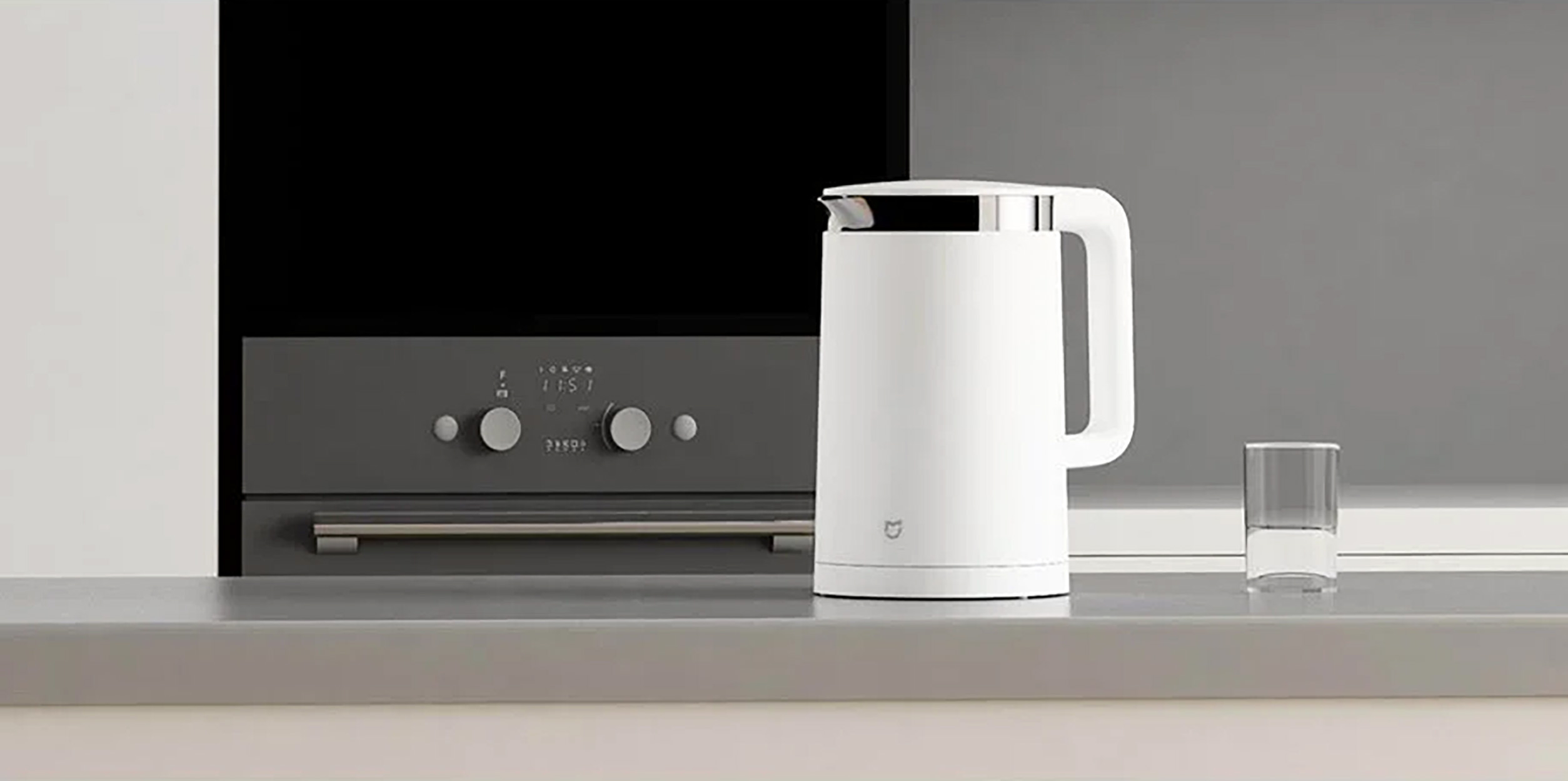 Witte elektrische waterkoker met zwarte voet en handvat. Geplaatst op een grijs oppervlak naast een glas water.
