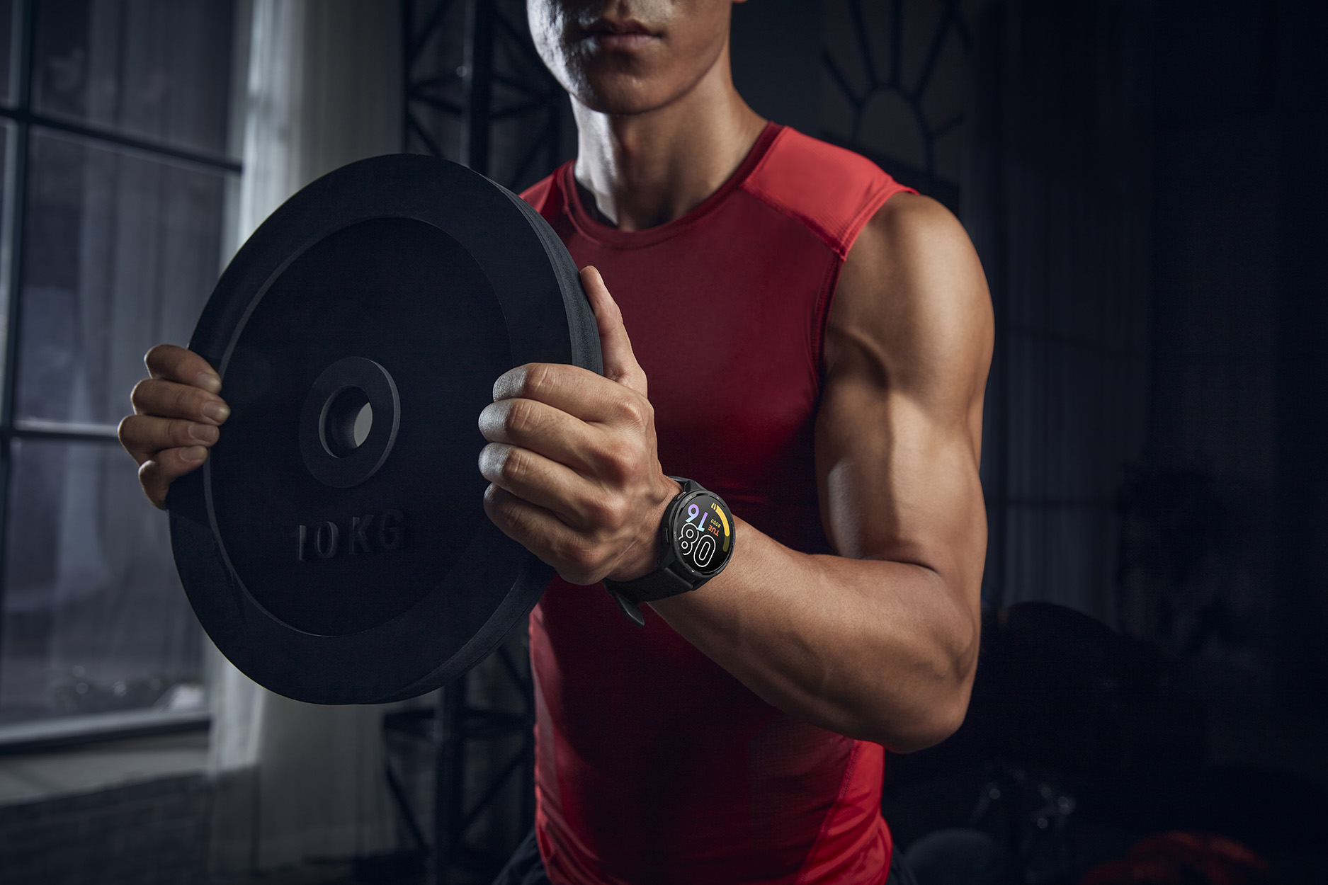 Ein Mann in einem roten Tanktop hält eine schwarze Gewichtsscheibe, mit Smartwatch am Handgelenk.