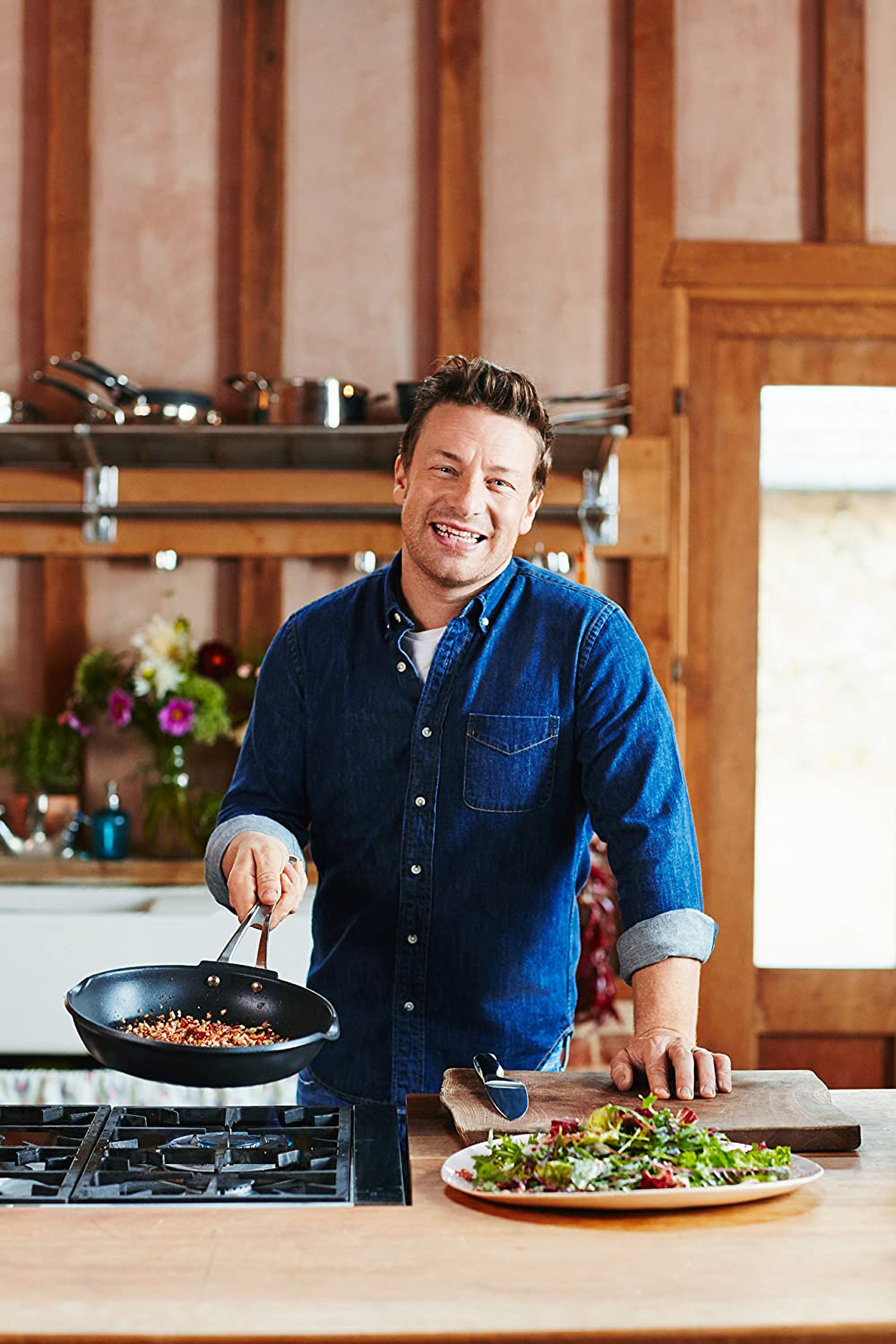 Homme dans la cuisine souriant, tenant une poêle avec de la nourriture. Fond en bois.