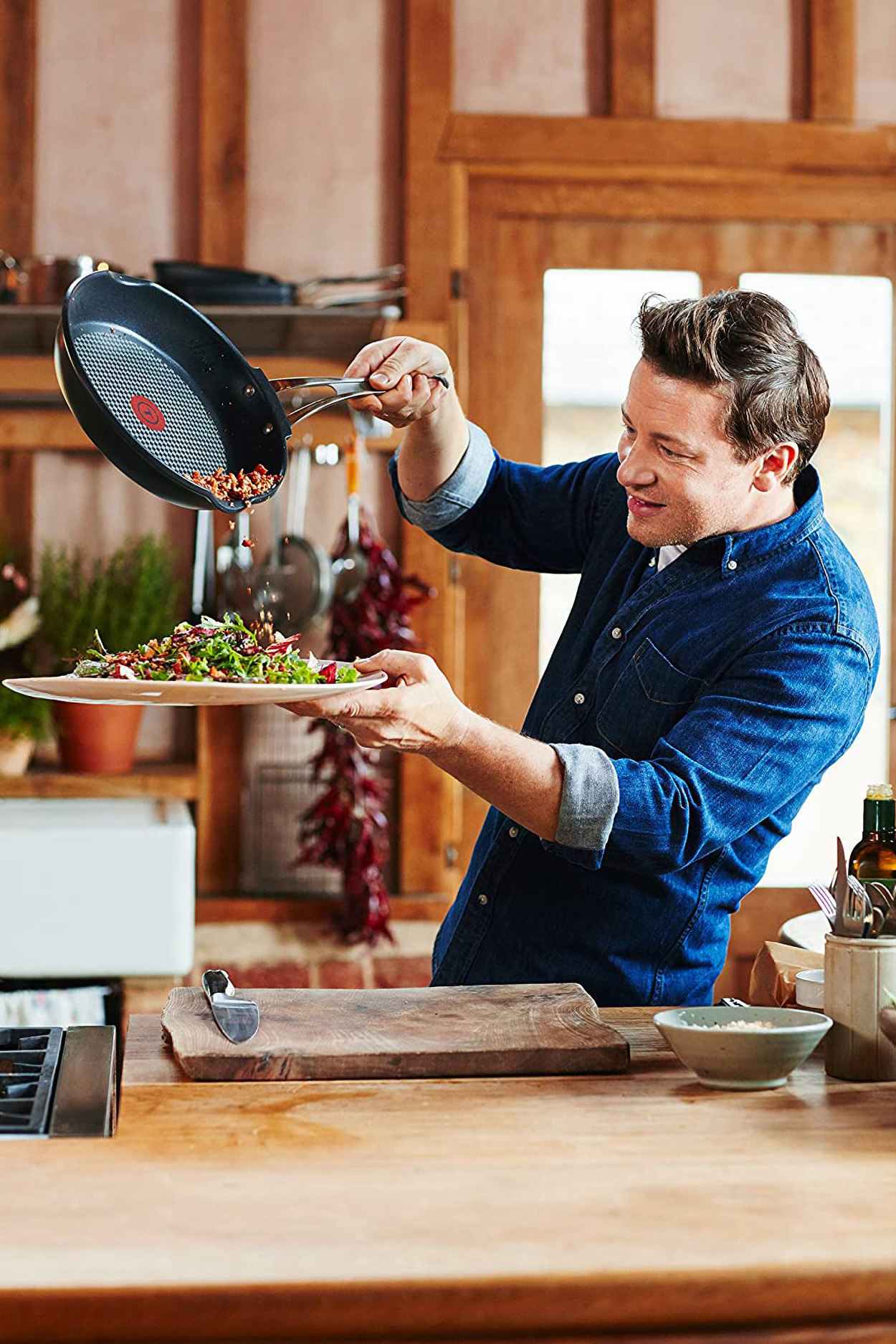 Chef en chemise bleue versant de la nourriture d'une poêle sur une assiette avec une salade, cuisine en arrière-plan.