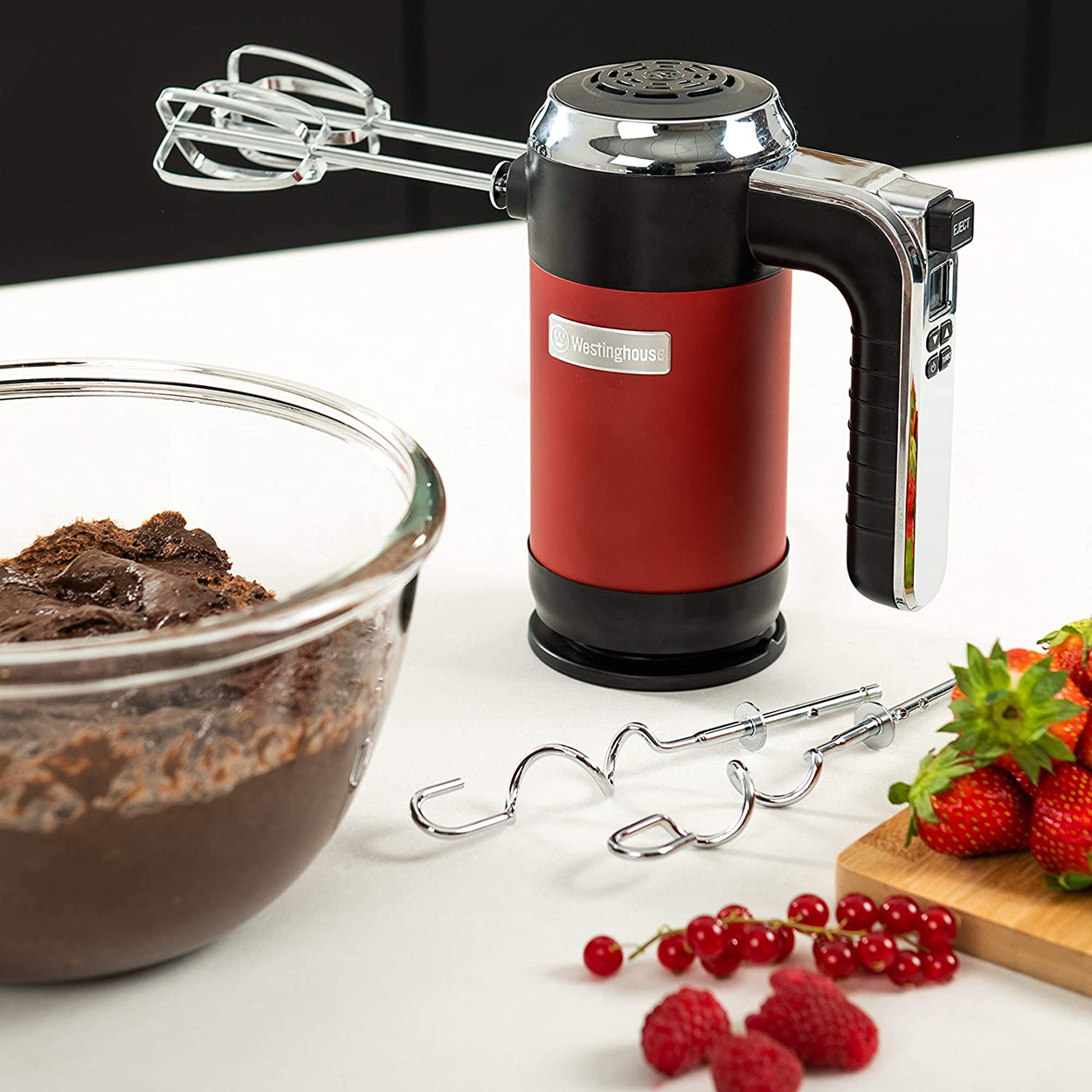Roter und schwarzer Handmixer mit Aufsätzen, Schüssel mit braunem Teig, Erdbeeren und Beeren auf weißer Oberfläche.