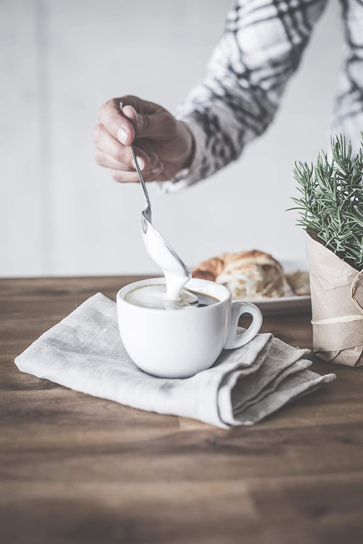 Eine Hand rührt Sahne in eine Kaffeetasse auf einem Holztisch, mit Croissant und Pflanze.