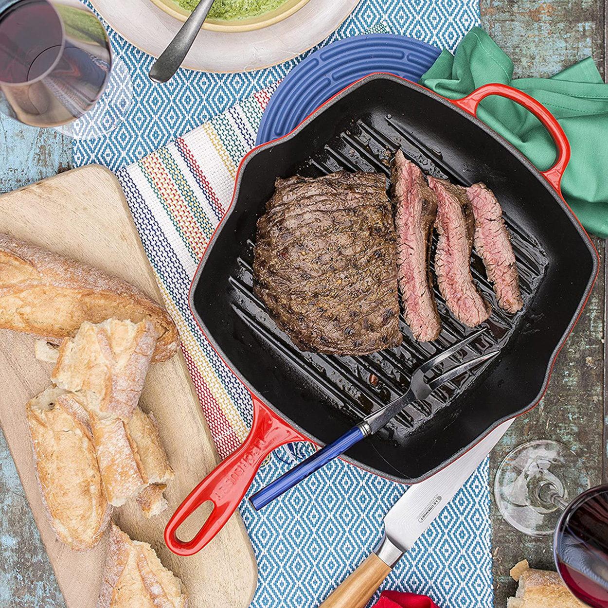 Vue aérienne d'un steak avec marques de grill, tranché, dans une poêle avec poignées rouges. D'autres aliments et boissons sont présents.