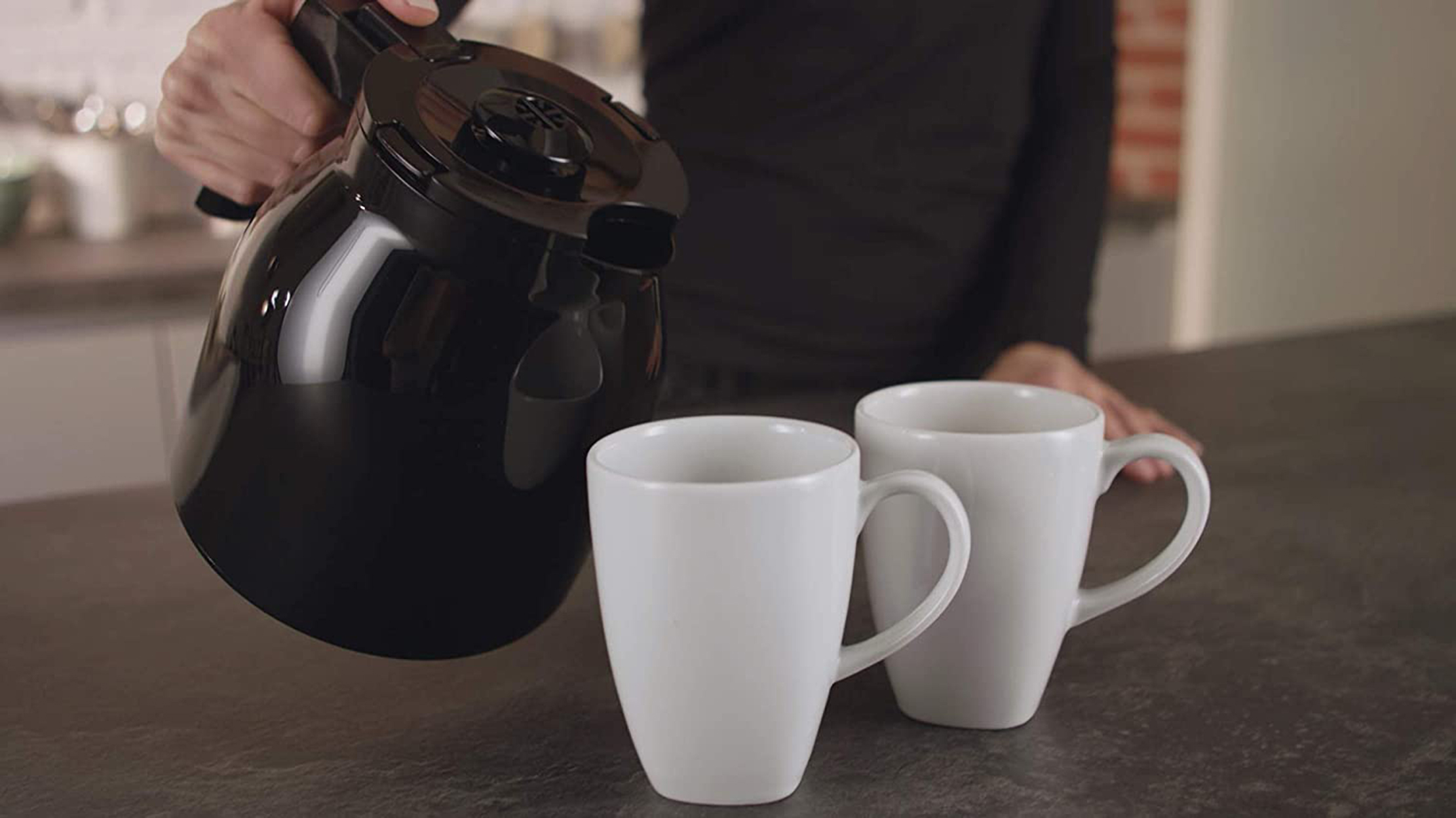 Osoba nalewa kawę z czarnego dzbanka do dwóch białych kubków. Tło kuchni.