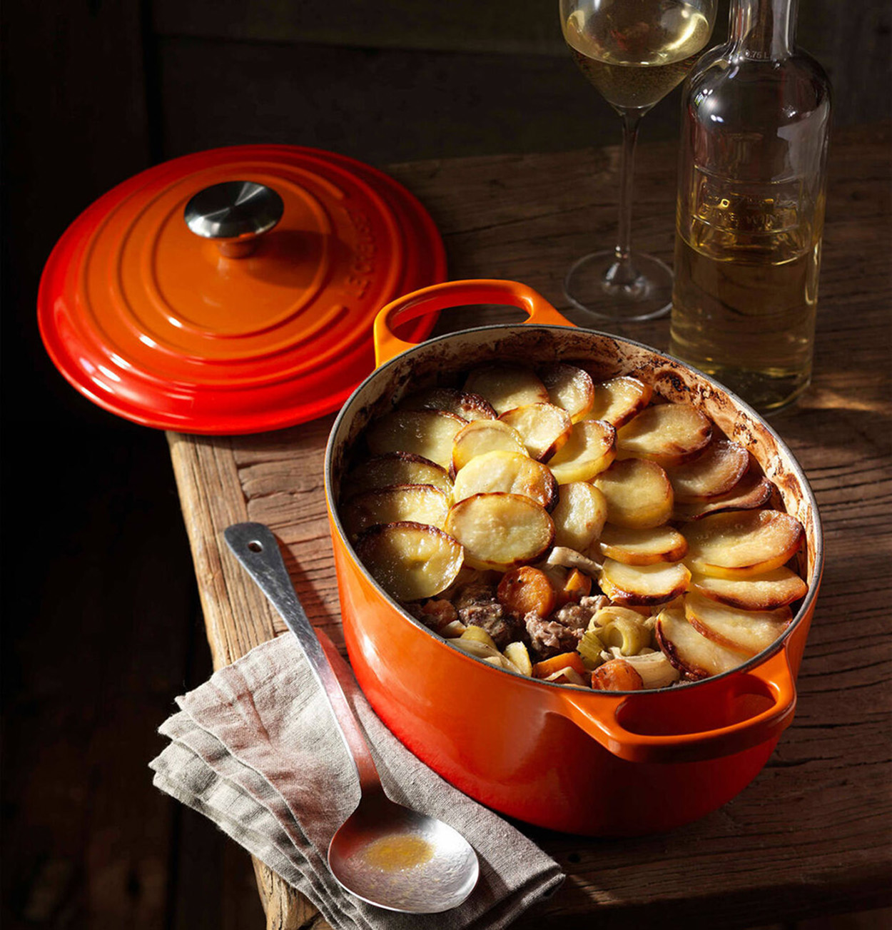Pomarańczowy garnek z ziemniakami, gulaszem i pokrywką. Kieliszek wina i butelka na drewnianym stole.