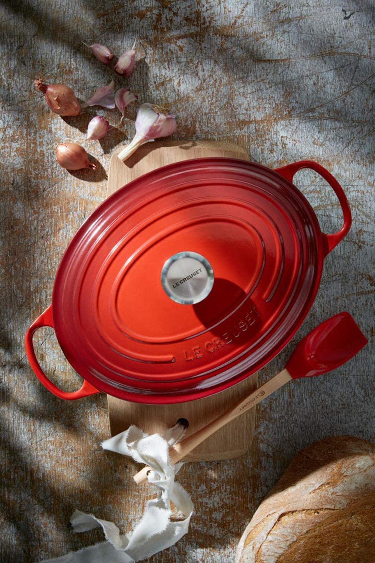 Roter Le Creuset ovaler Bräter mit Deckel, auf einem Holzbrett, mit Knoblauch und Schalotten.