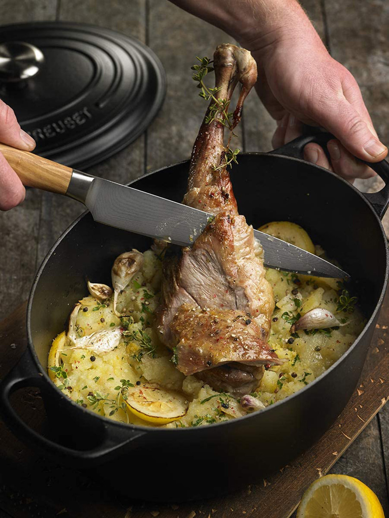 Un gigot d'agneau rôti dans une casserole ovale noire, coupé avec un couteau par les mains d'une personne.