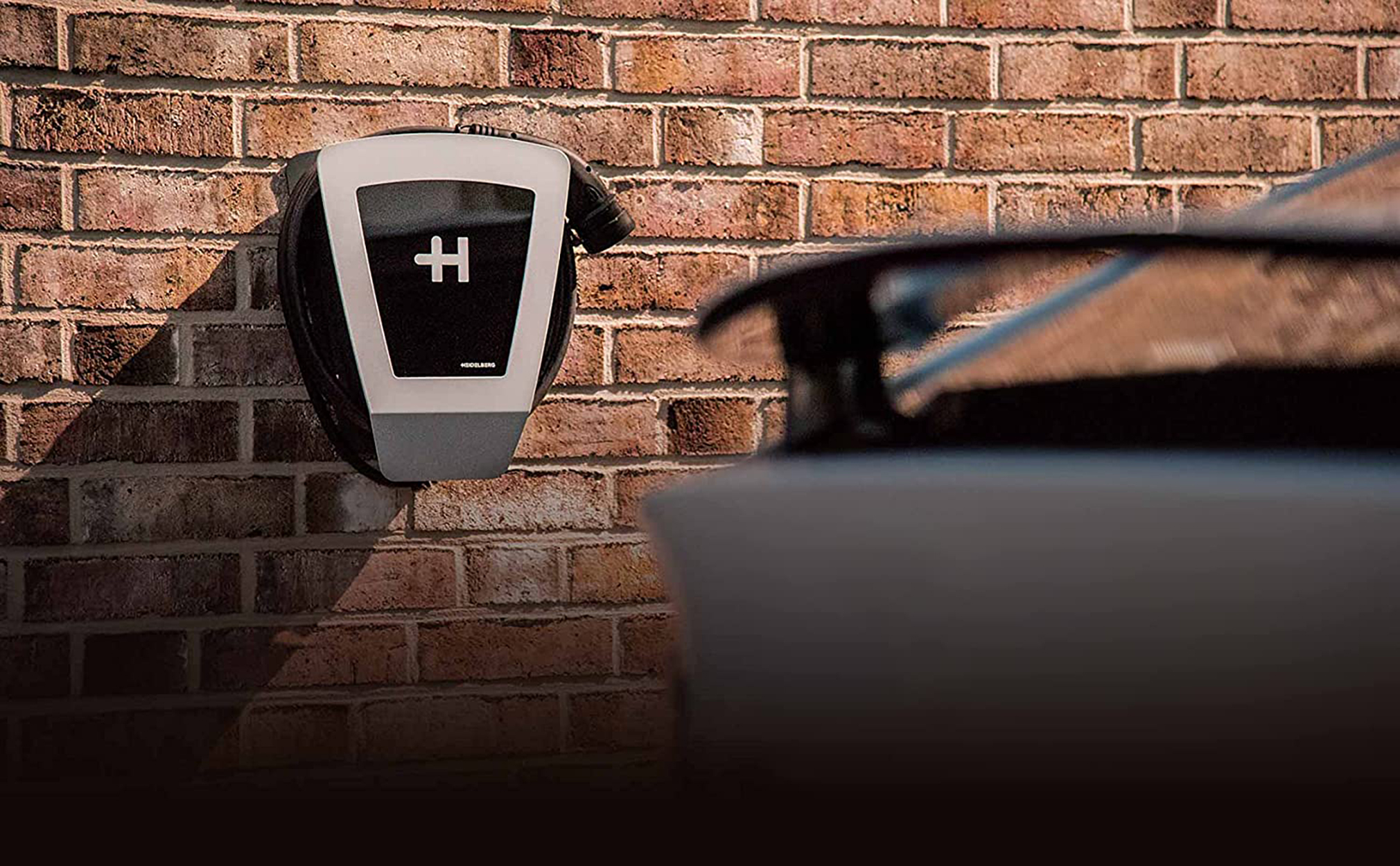 Elektroauto-Ladegerät an einer Ziegelmauer montiert, Auto teilweise sichtbar.