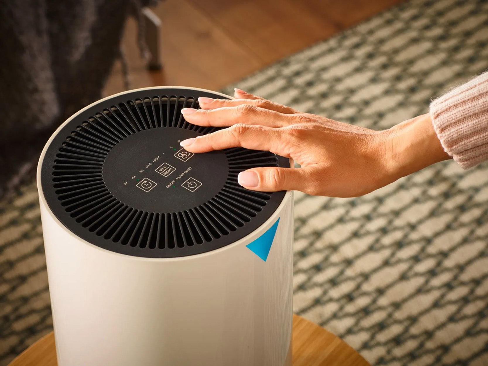 Eine Hand berührt das Bedienfeld eines weißen Luftreinigers, der auf einem Holztisch steht, mit einem Teppich im Hintergrund.