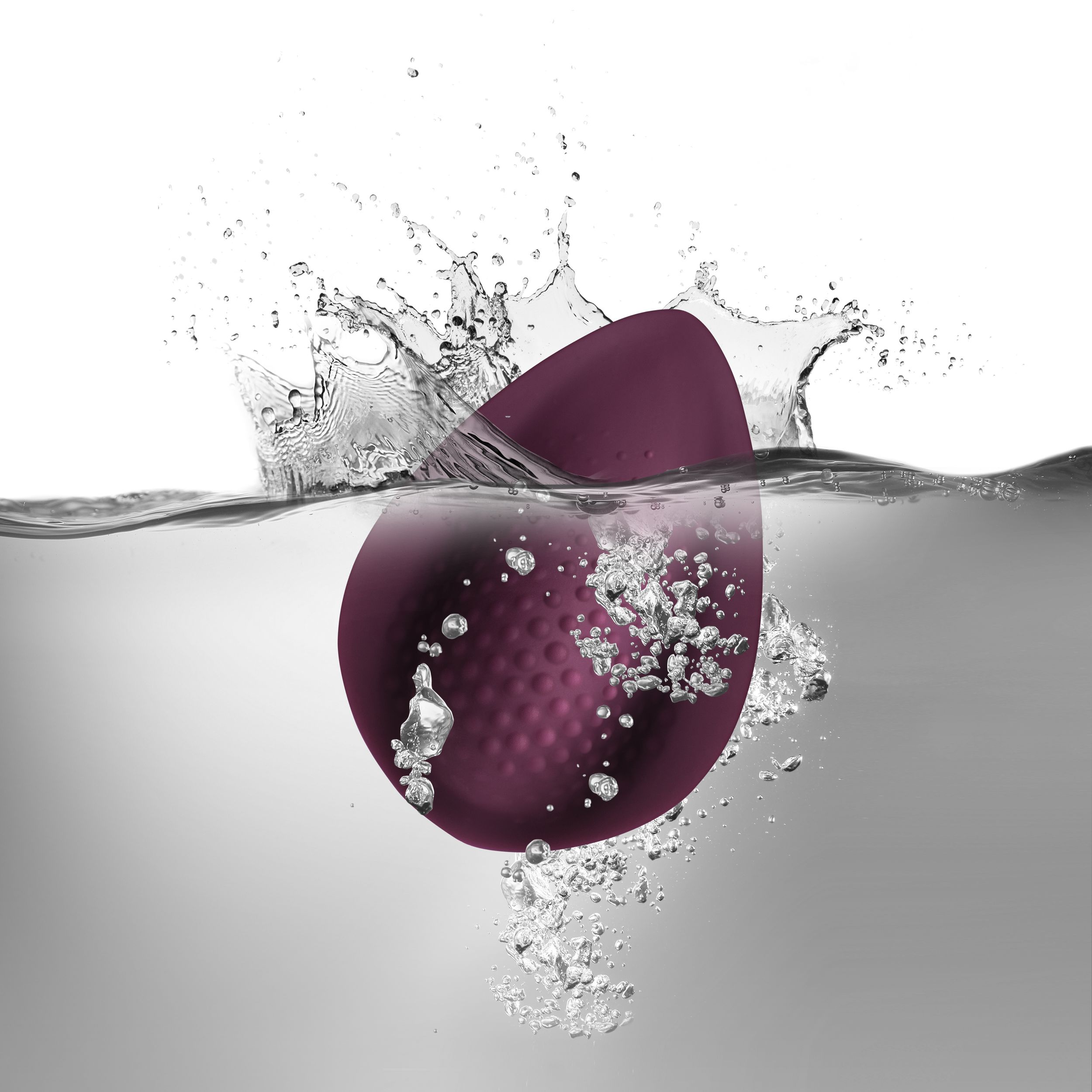 Ein lila Sexspielzeug, das in Wasser getaucht ist und Blasen und Spritzer erzeugt. Der Hintergrund ist grau.