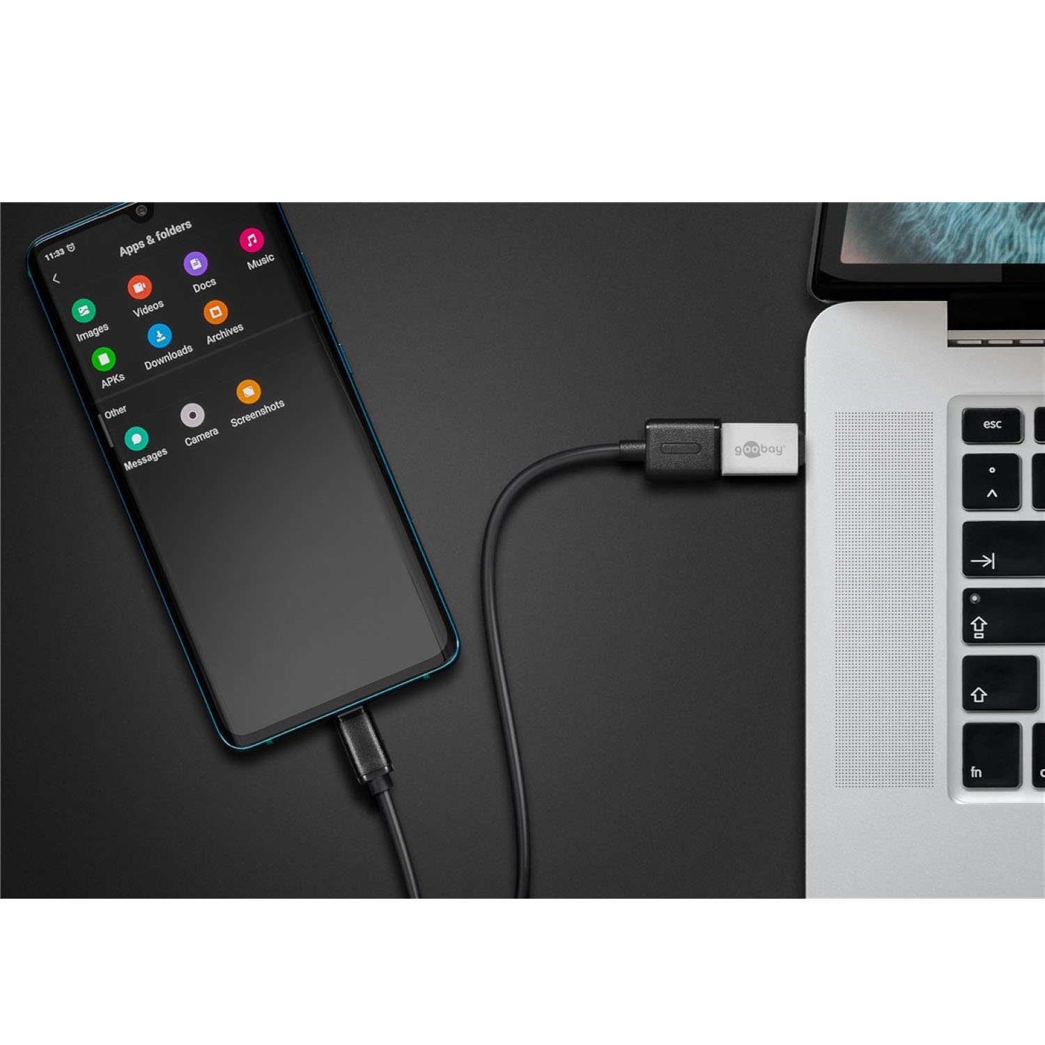 Telefon jest podłączony do laptopa za pomocą czarnego kabla. Telefon wyświetla ikony aplikacji. Biała powierzchnia.