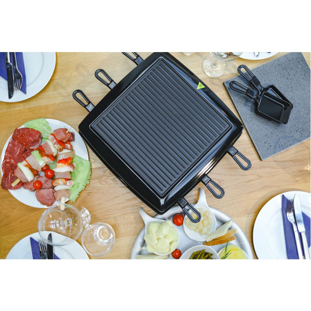Raclette-Grill mit Essen auf Tellern, einschließlich Fleisch, Spießen und Gemüse, auf Holztisch.