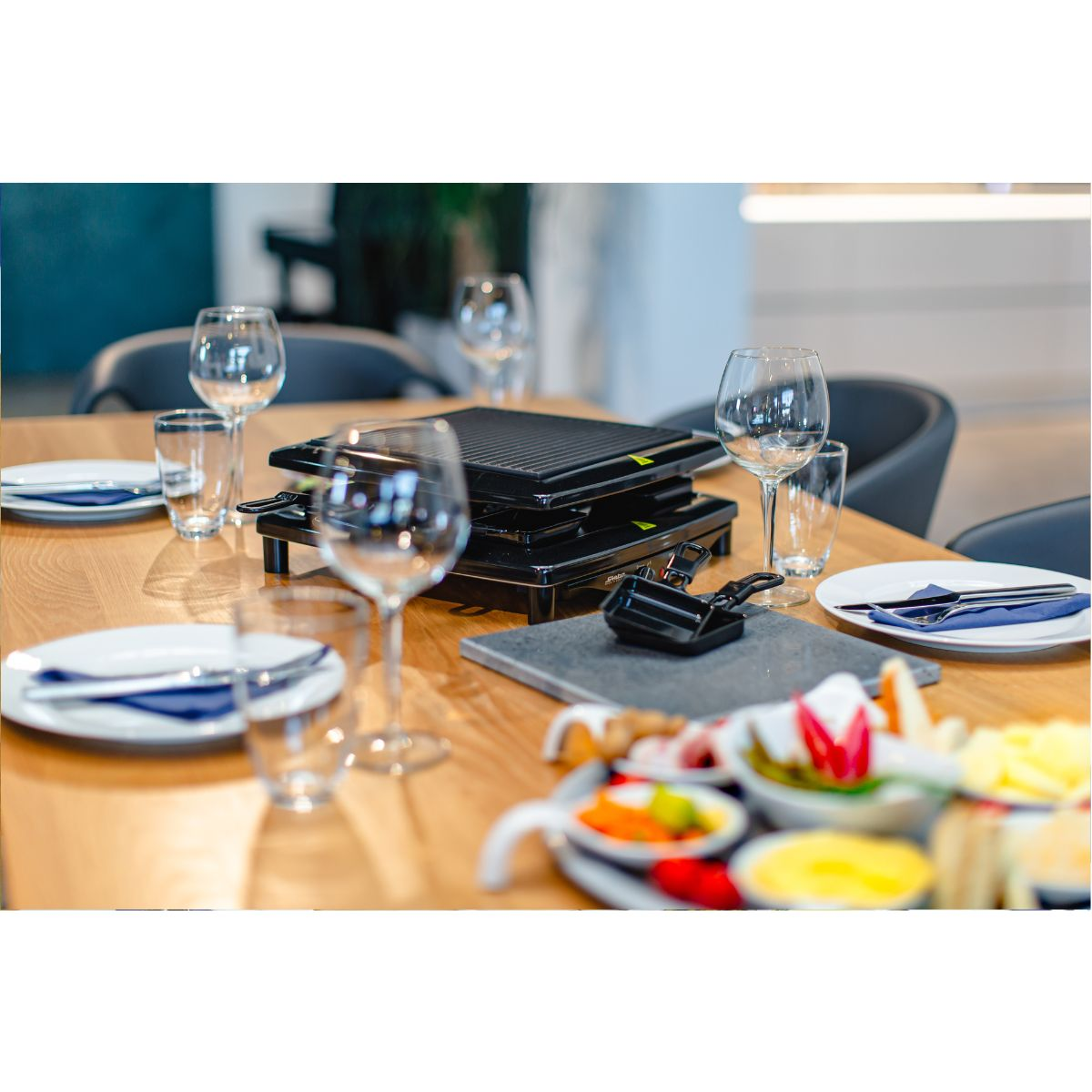 Ein Tischgedeck mit Gläsern, einem Raclette-Grill und verschiedenen Lebensmitteln.