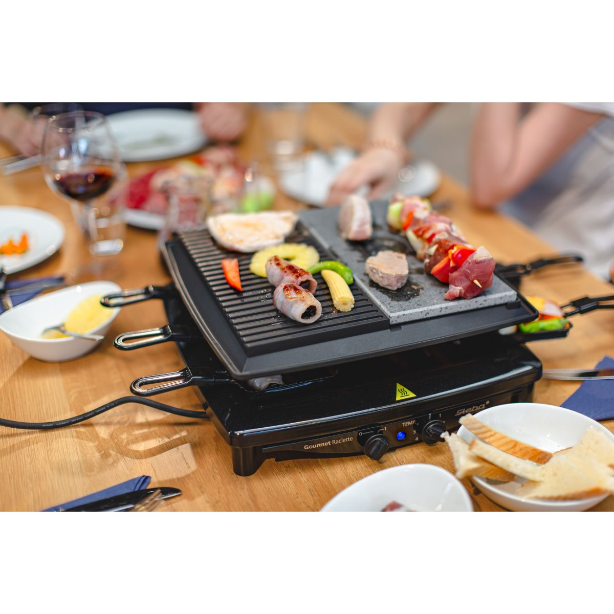 Raclette-Grill mit verschiedenen Speisen. Hände von Personen im Hintergrund.