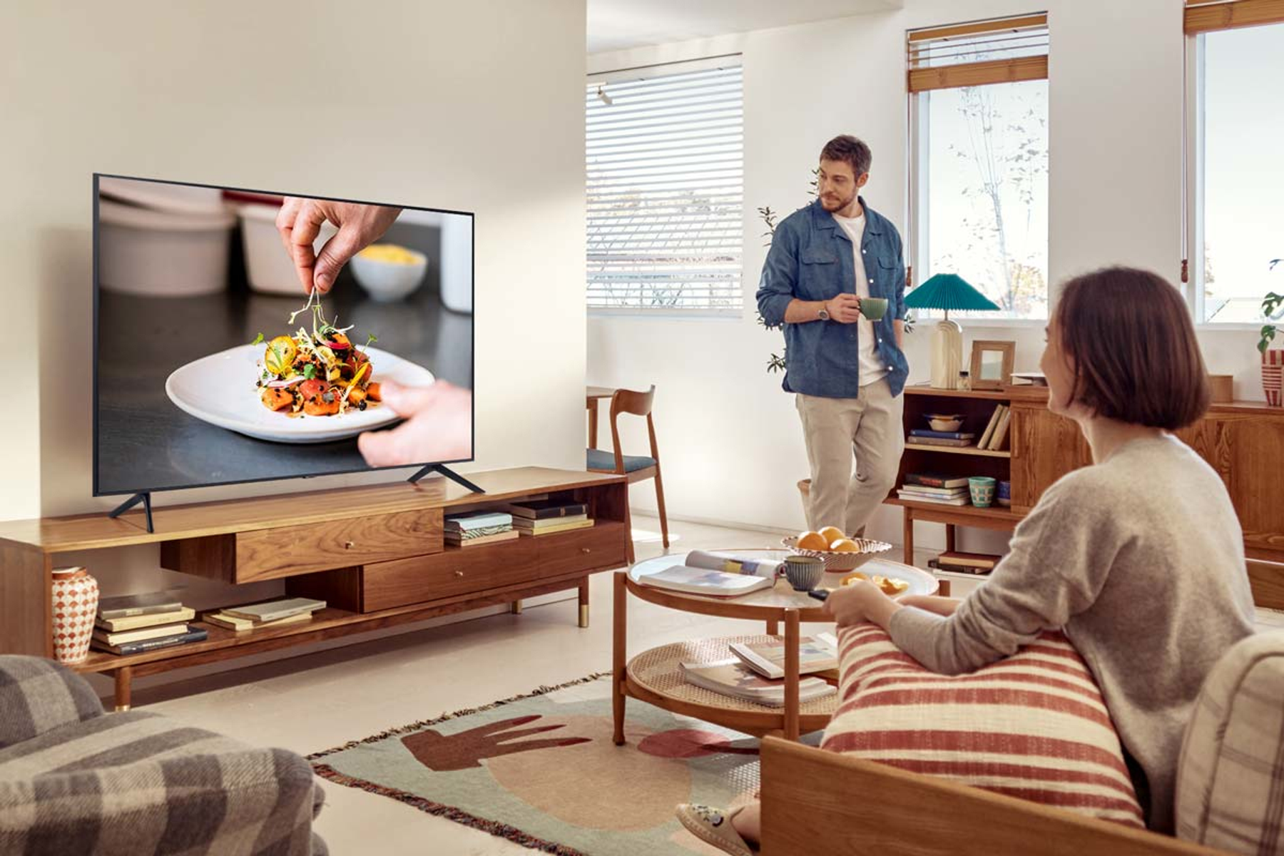 Una TV mostra cibo su un piatto. Un uomo e una donna sono in un soggiorno. La stanza è luminosa.