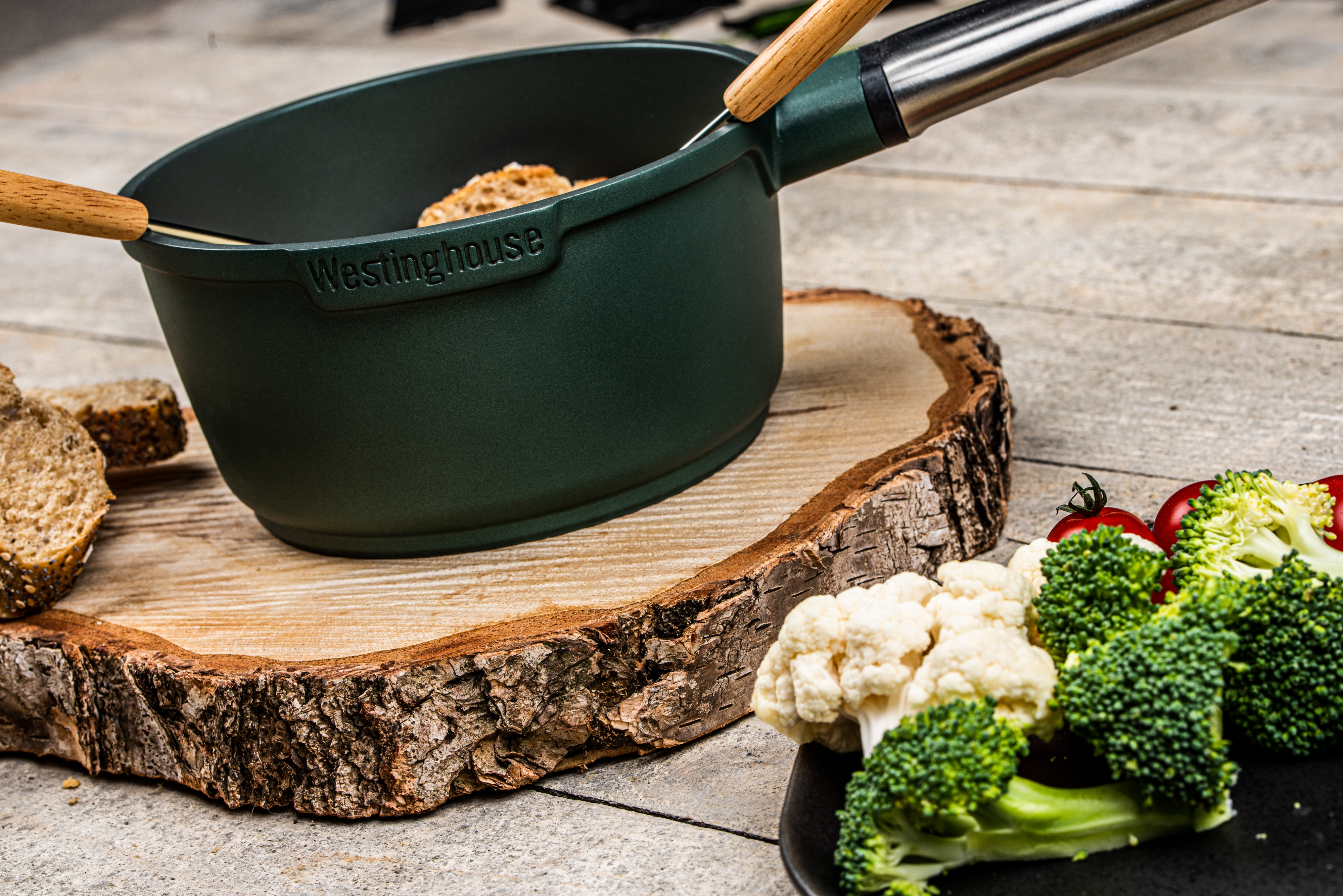 Ein dunkelgrüner Fondue-Topf mit Holzgriffen, auf Holzuntergrund mit Lebensmitteln.