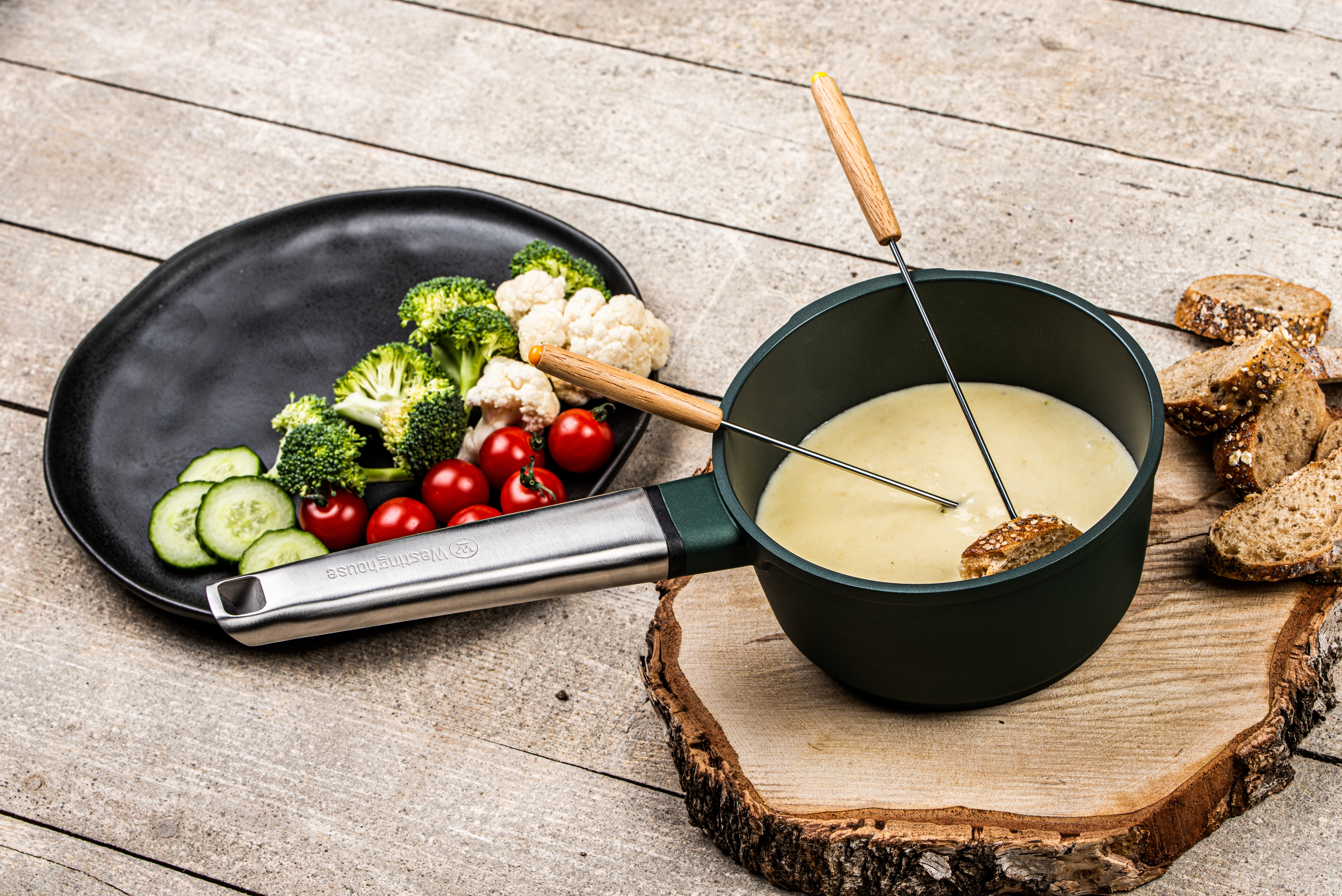 Ein Fonduetopf mit Käse und Brot. Brokkoli, Tomaten und Gurken auf einem Teller.
