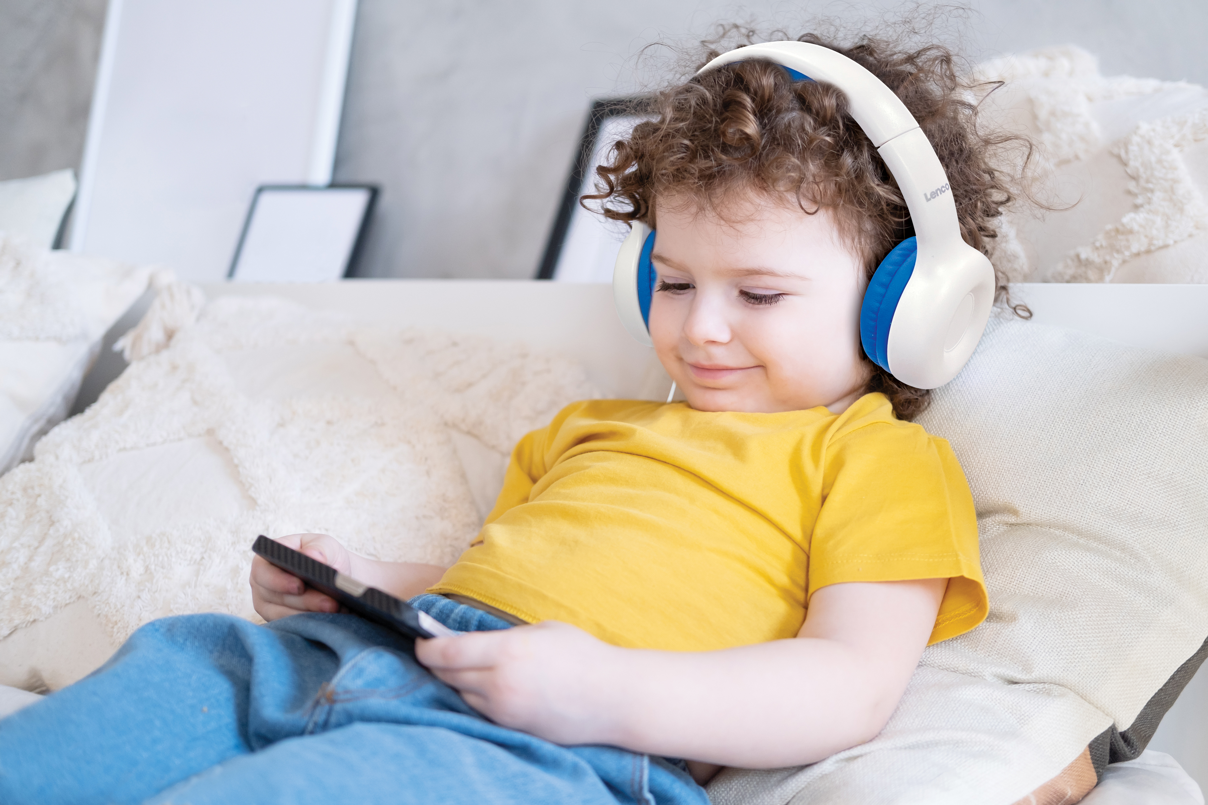 Dziecko w białych i niebieskich słuchawkach, uśmiechnięte, trzymające tablet na sofie.
