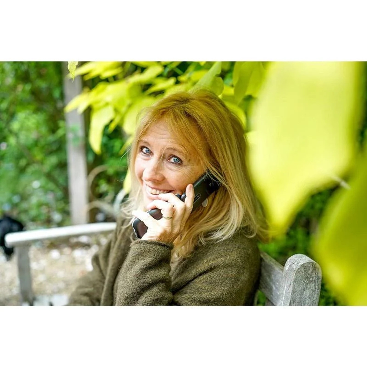 Kobieta z blond włosami uśmiecha się, trzymając telefon przy uchu, siedząc na ławce na zewnątrz.