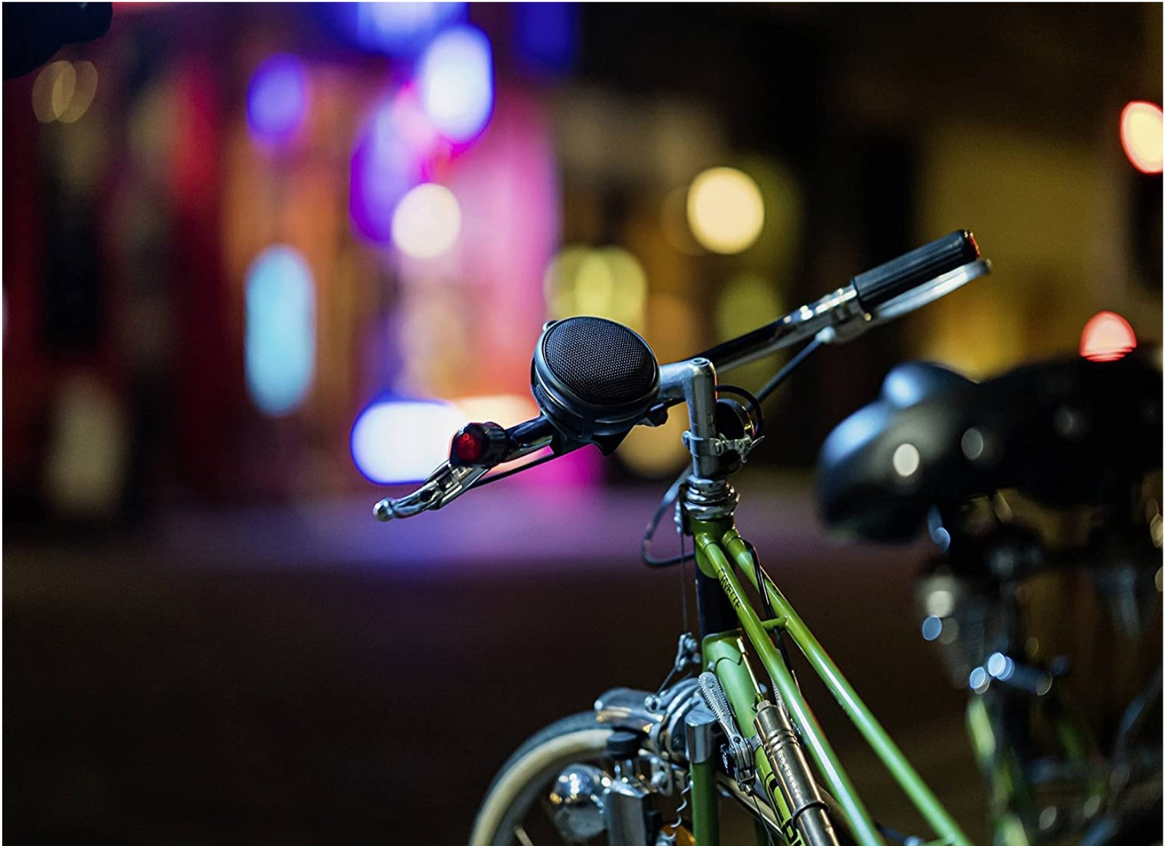 Ein grüner Fahrradlenker mit Klingel, Lichtern und verschwommenem, farbenfrohem Stadt-Hintergrund.