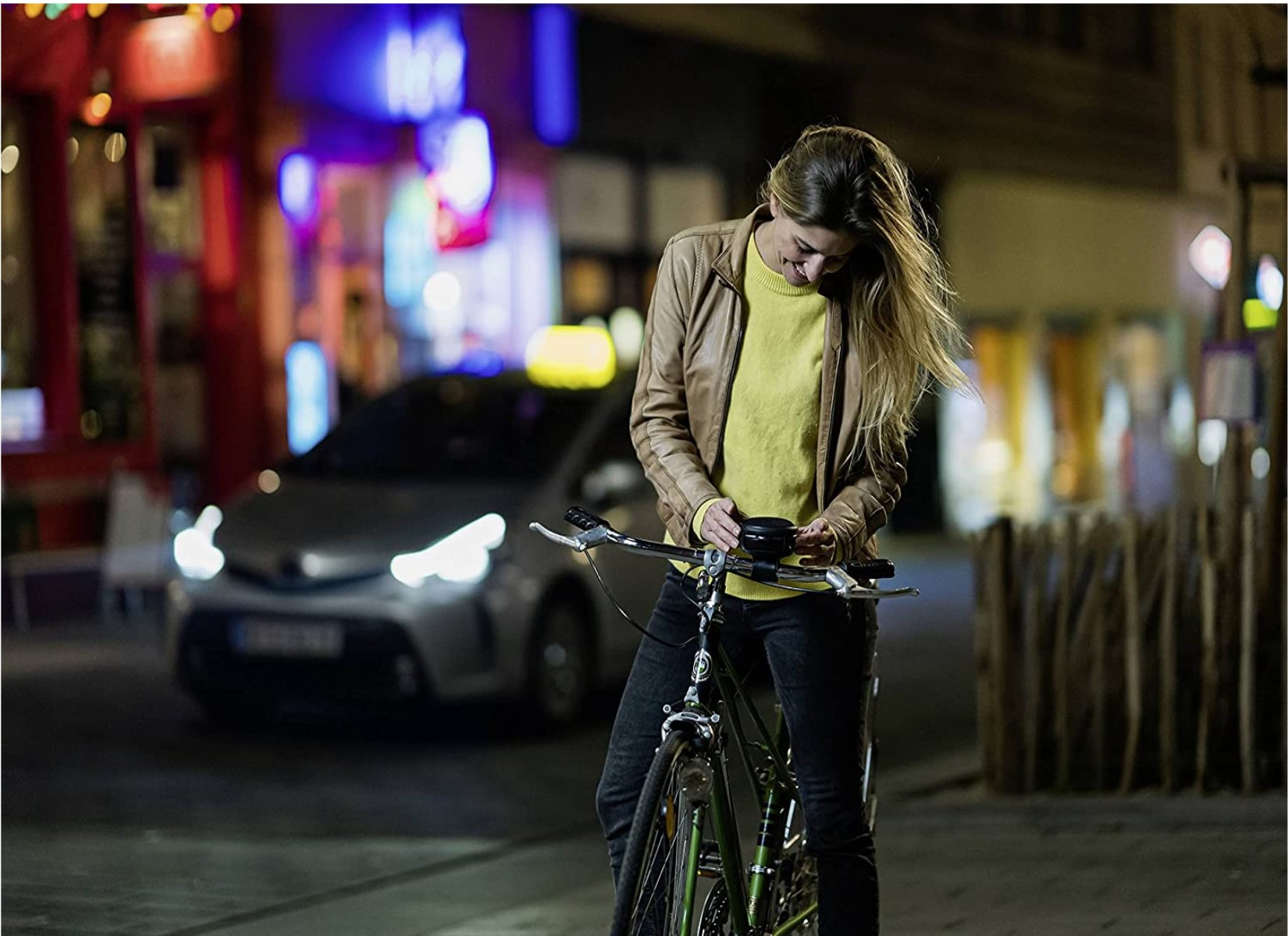 Frau auf einem Fahrrad bei Nacht, schaut auf ihr Handy. Auto und Stadtlichter im Hintergrund.