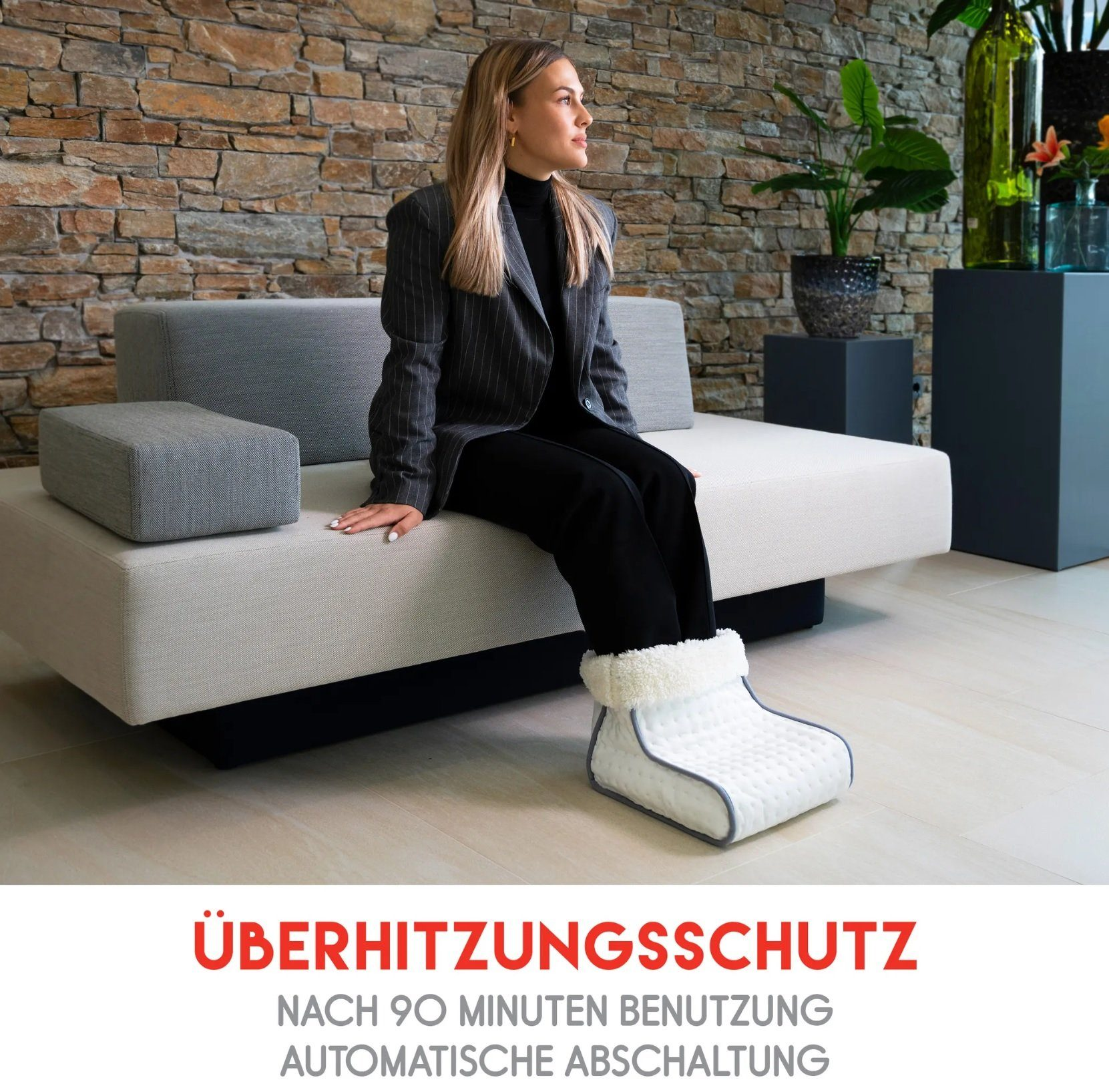 Eine Frau sitzt auf einem Sofa und trägt einen Blazer und einen Fußwärmer. Text auf Deutsch über Überhitzungsschutz.